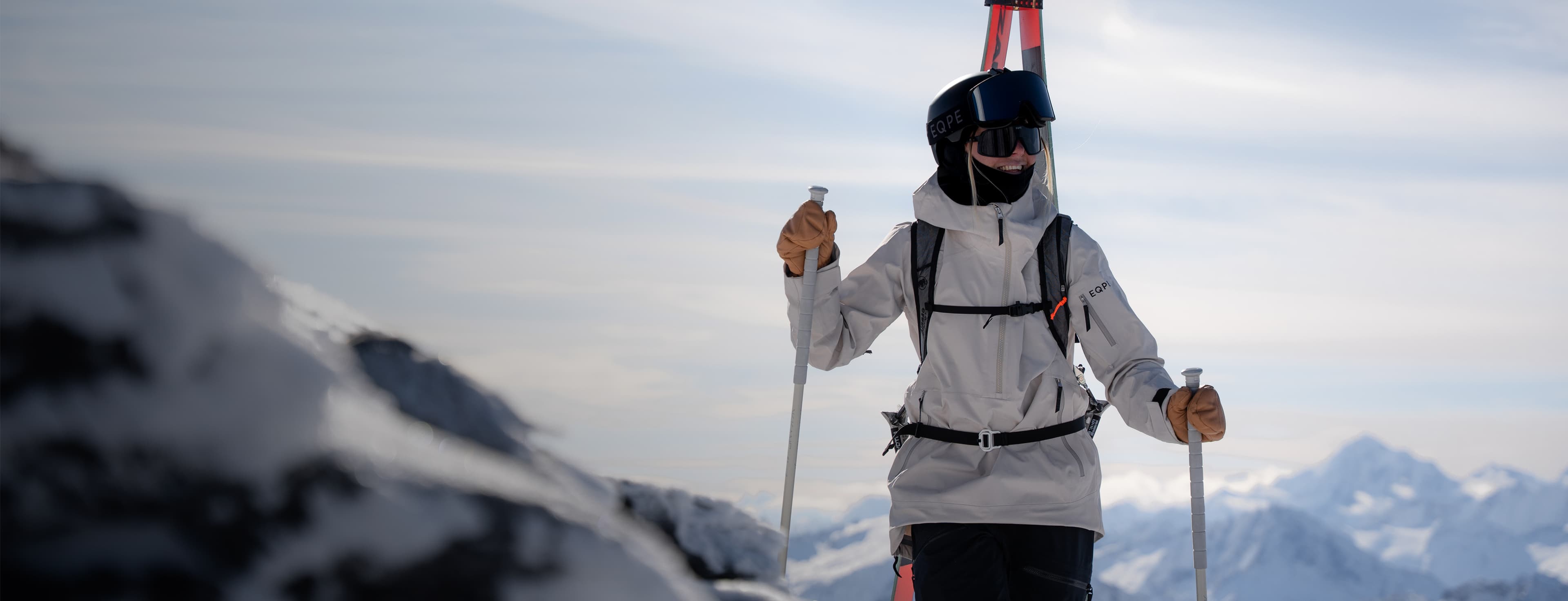 A skier in a beige jacket and dark pants stands on a snowy mountain, holding poles, with snow-covered peaks in the background.