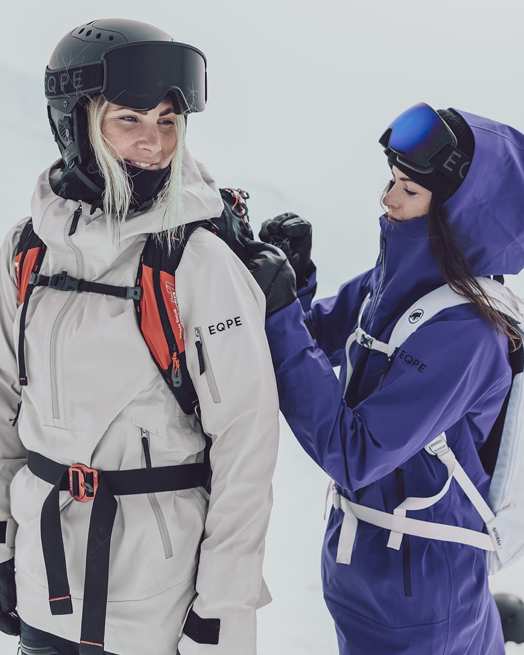 Two people in winter clothes, one in beige and one in purple. One is adjusting the other's backpack.