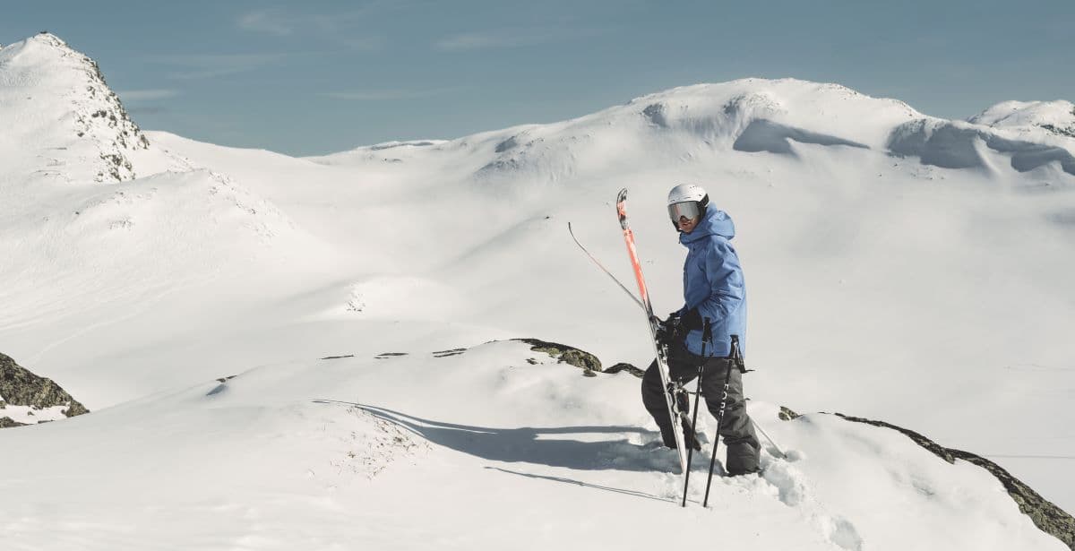 A skier in a blue jacket stands on a snowy mountain peak, holding skis and poles, with expansive snow-covered mountains in the background.