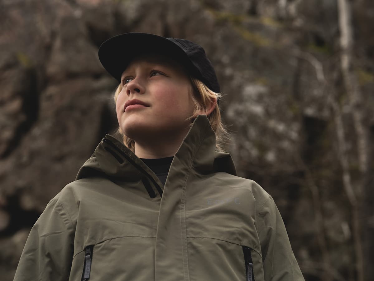 Young person in a green jacket and black cap, looking upward, with a rocky background.