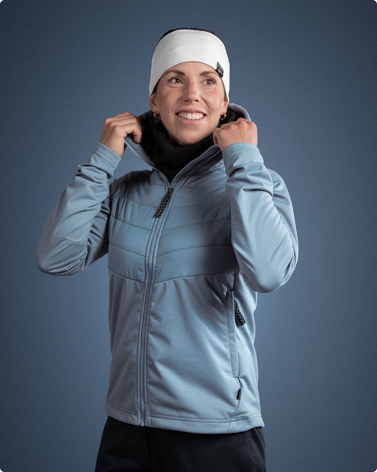 Person in a blue jacket and white headband smiling, adjusting their collar against a plain blue background.