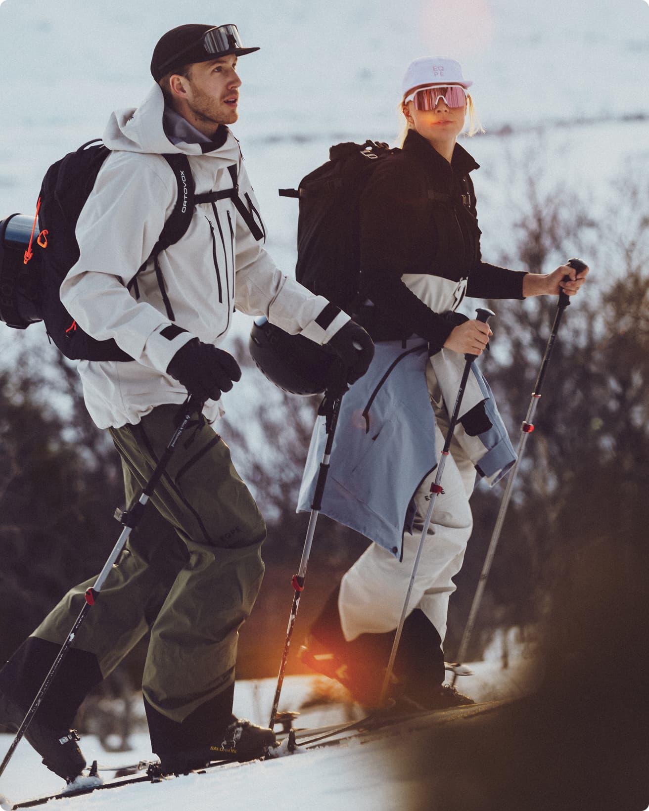 Two people in winter gear skiing uphill with poles, wearing backpacks and sunglasses, against a snowy landscape with sunlight in the background.