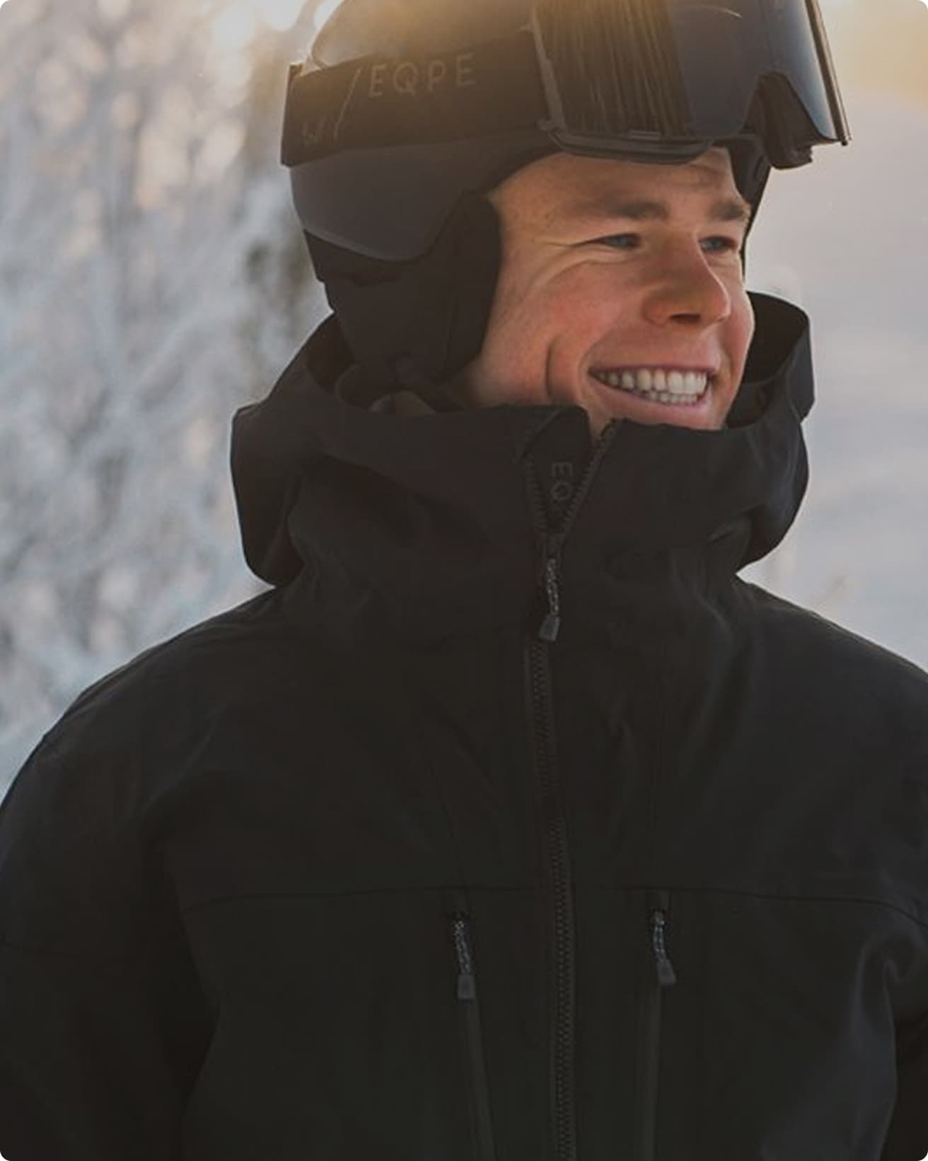 Person in a black jacket and helmet smiles outdoors, with snow-covered trees in the background and a warm glow from the sun.