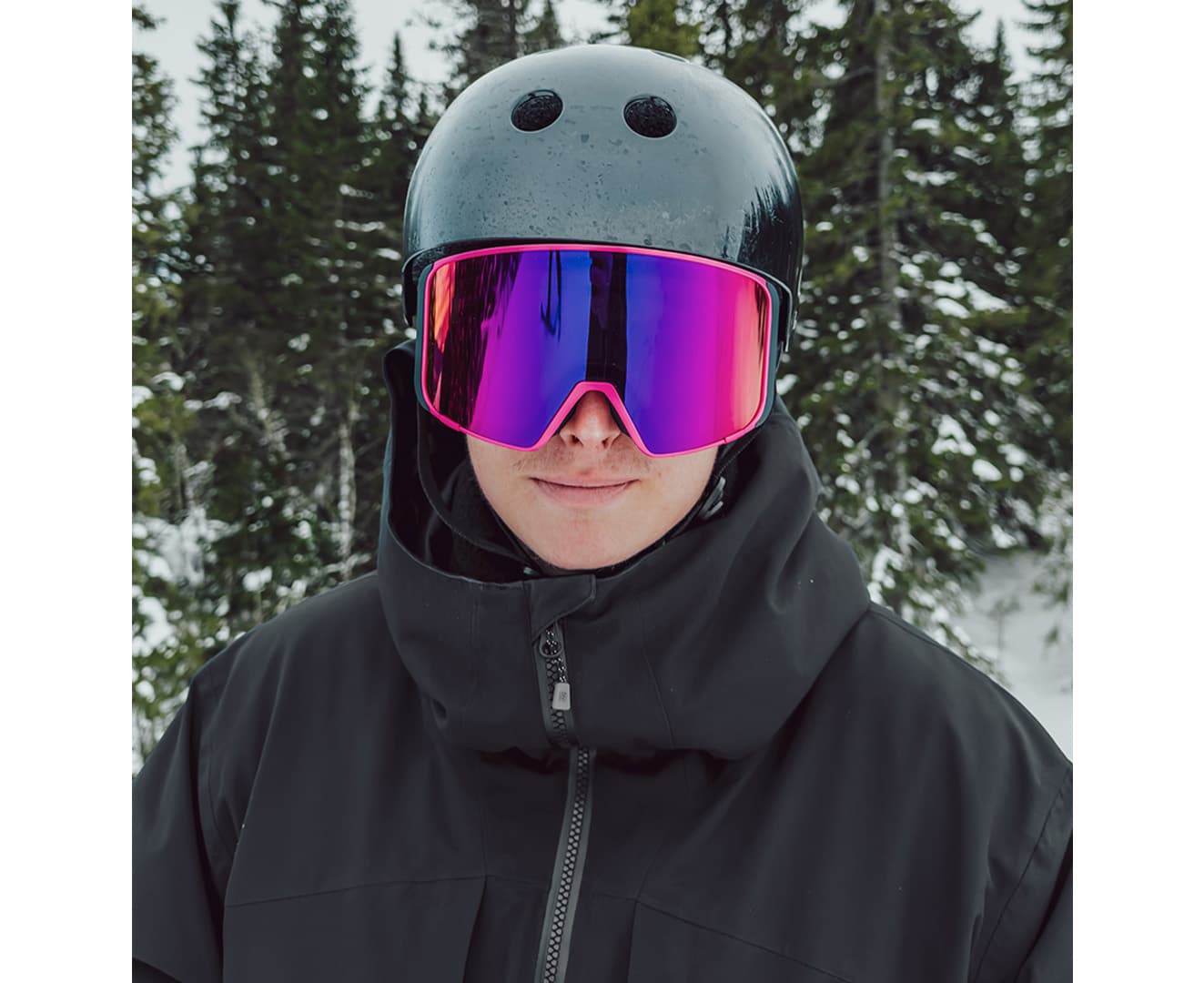 Oliwer wearing a black helmet, reflective ski goggles, and a black jacket stands outdoors with snow and trees in the background.