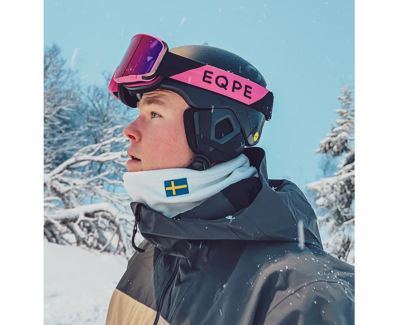 Walter in ski gear with pink goggles and a Swedish flag neck warmer stands in a snowy landscape, looking ahead.