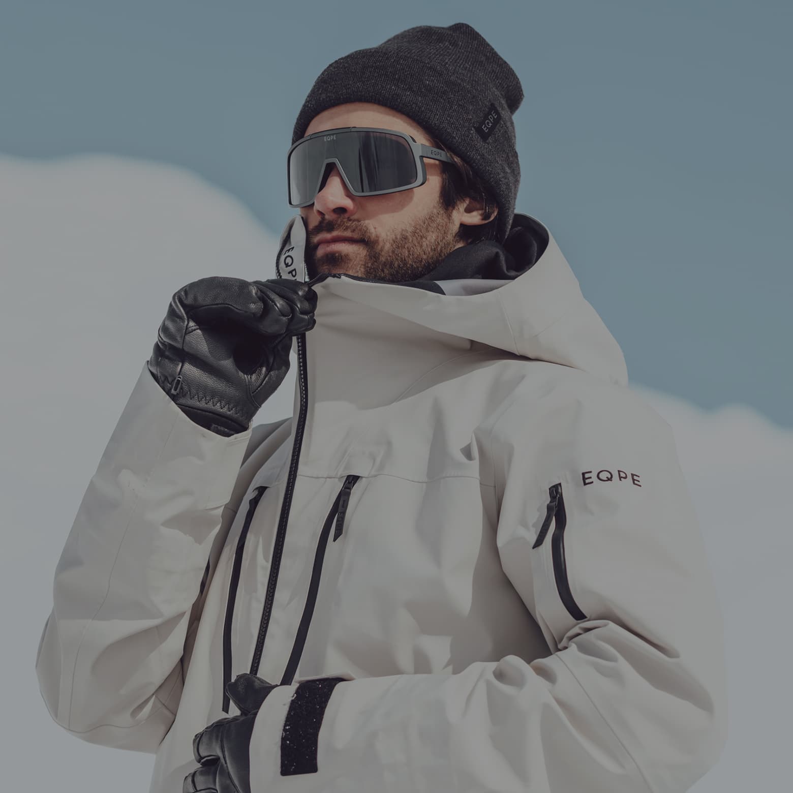 A person wearing a beige winter jacket, dark beanie, sunglasses, and gloves, zipping up their coat in a snowy outdoor setting.