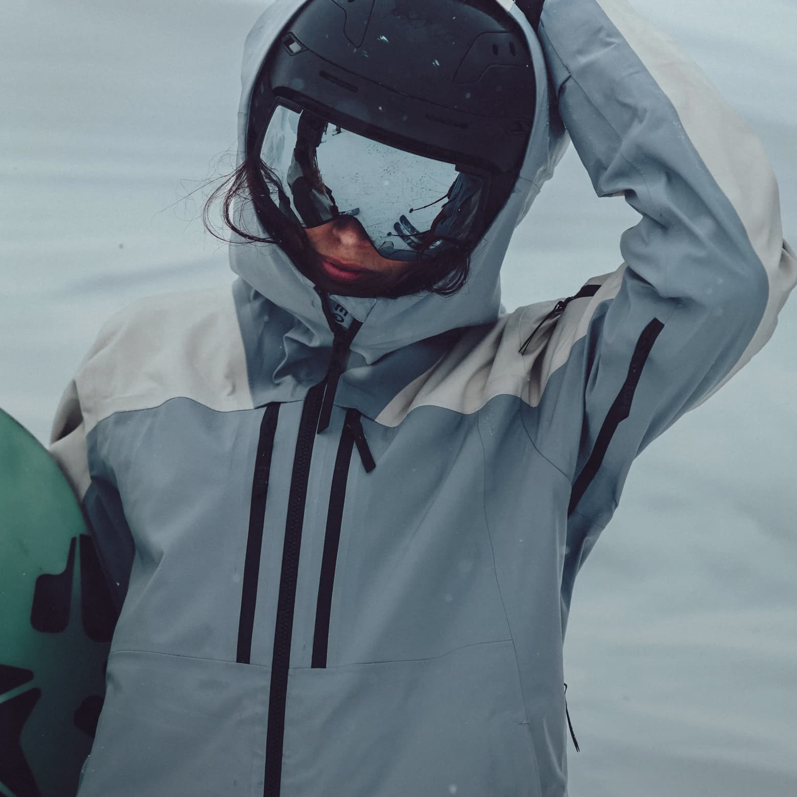 Person in a winter jacket and helmet holding a snowboard, standing in a snowy landscape, with goggles reflecting the surroundings.