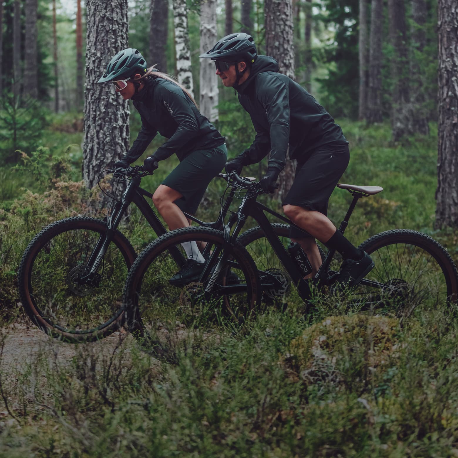 Two cyclists in dark gear ride mountain bikes through a lush, green forest trail.