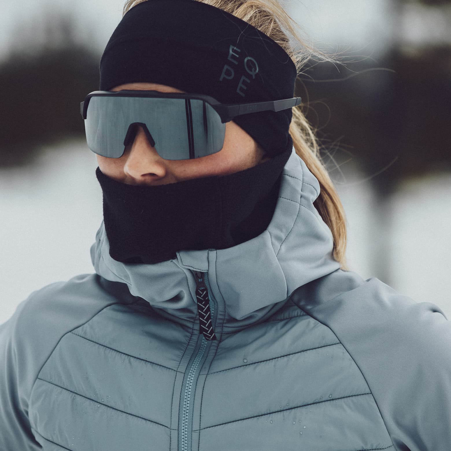 Person dressed in winter gear, including sunglasses and a headband, stands in a snowy landscape with blurred trees in the background.