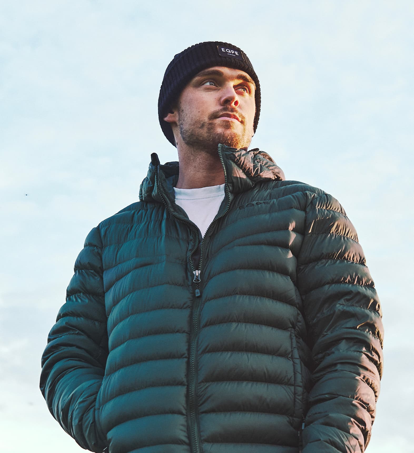 Man in a green liner jacket and black beanie looks into the distance.