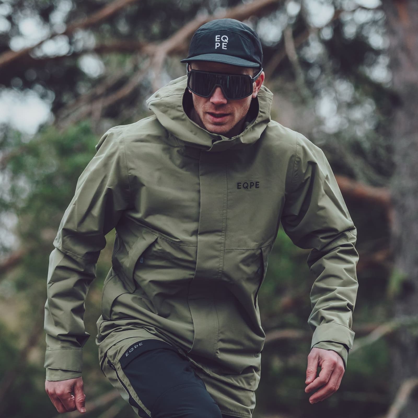 Person in a green jacket and black cap with sunglasses, outdoors in a forest setting.