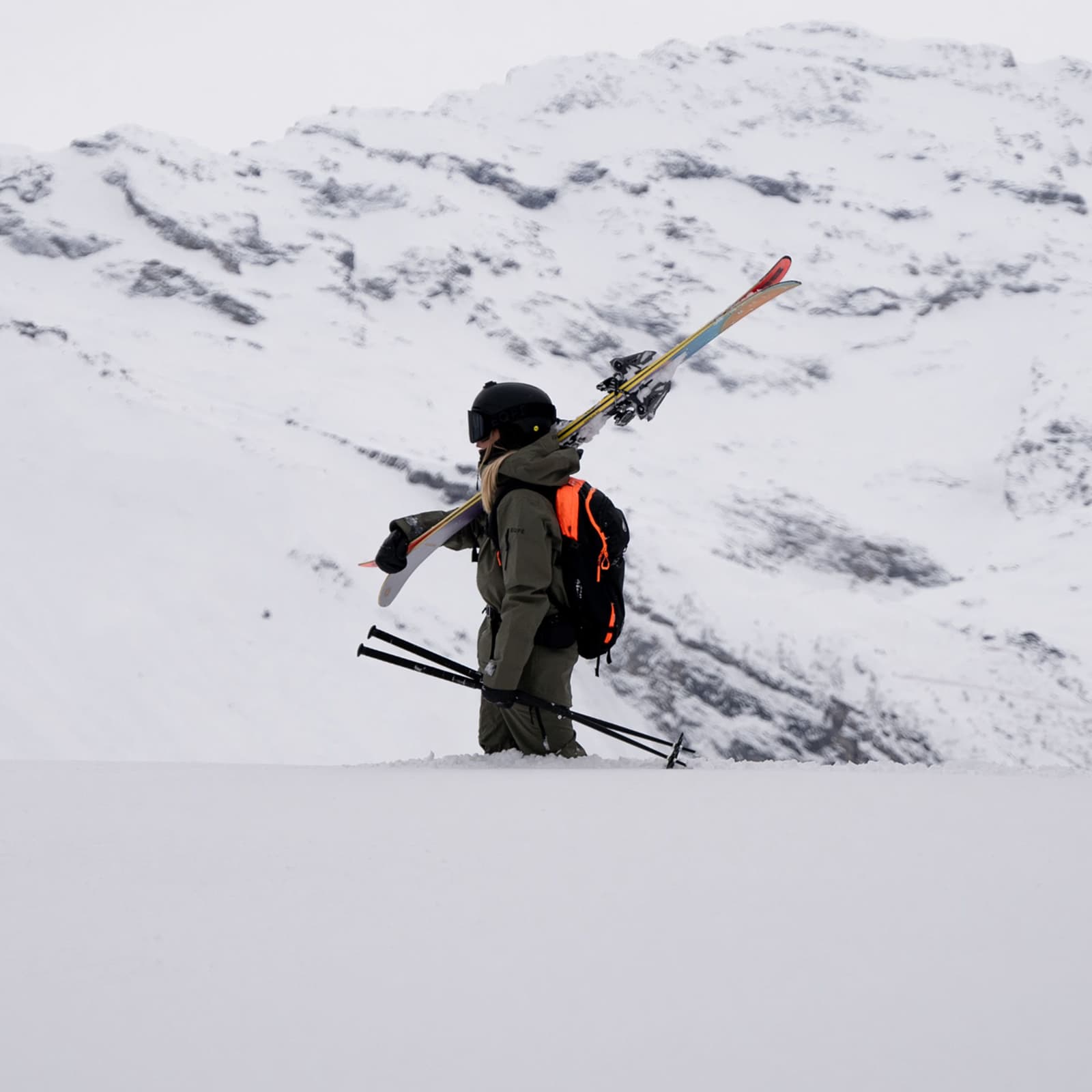 Skier in snowy landscape, carrying skis and poles on their shoulder, surrounded by snow-covered trees and mountains.