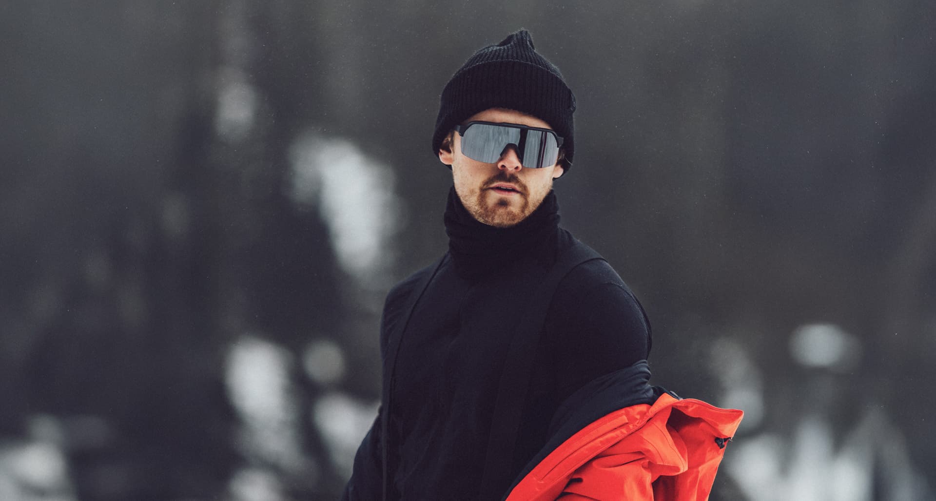 A man in a black beanie and sunglasses stands in a snowy landscape, wearing a black outfit with a red jacket draped over his shoulder.