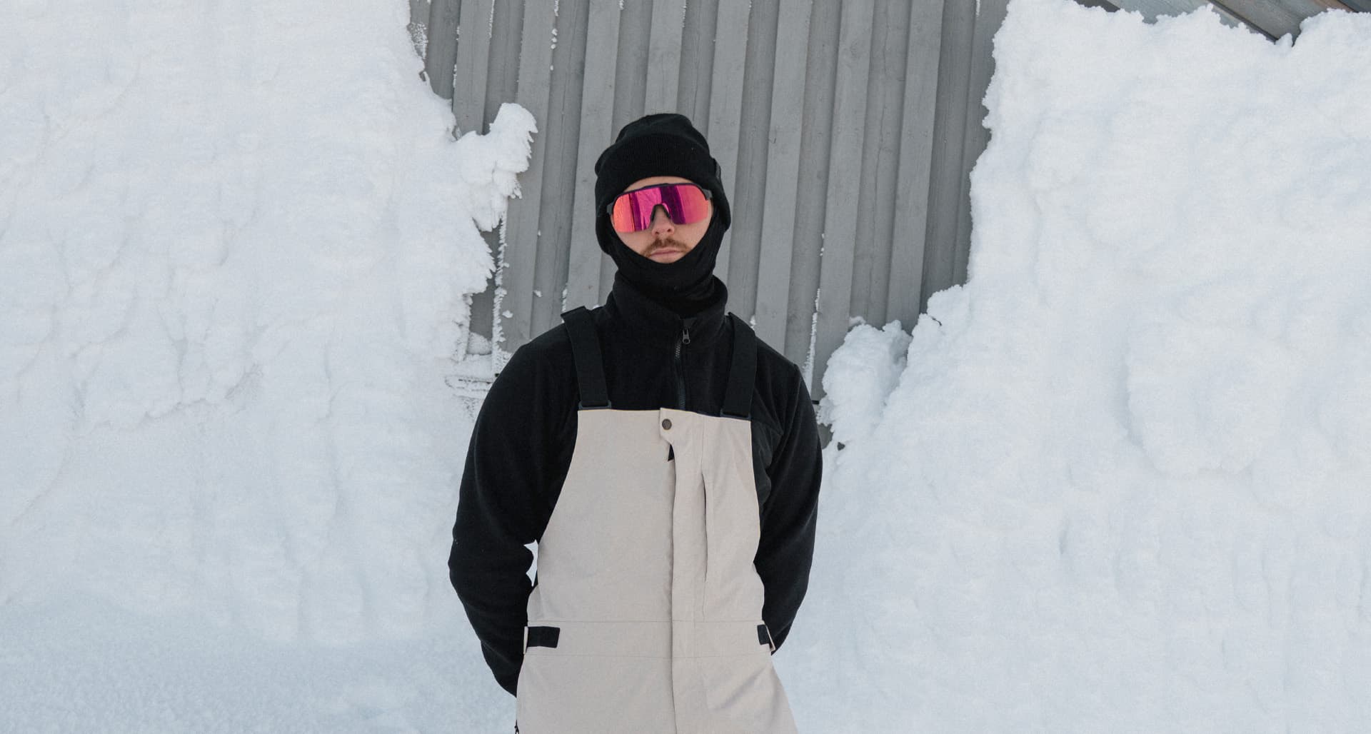 Person in snow gear with reflective goggles and a beanie stands in front of a snow-covered building.
