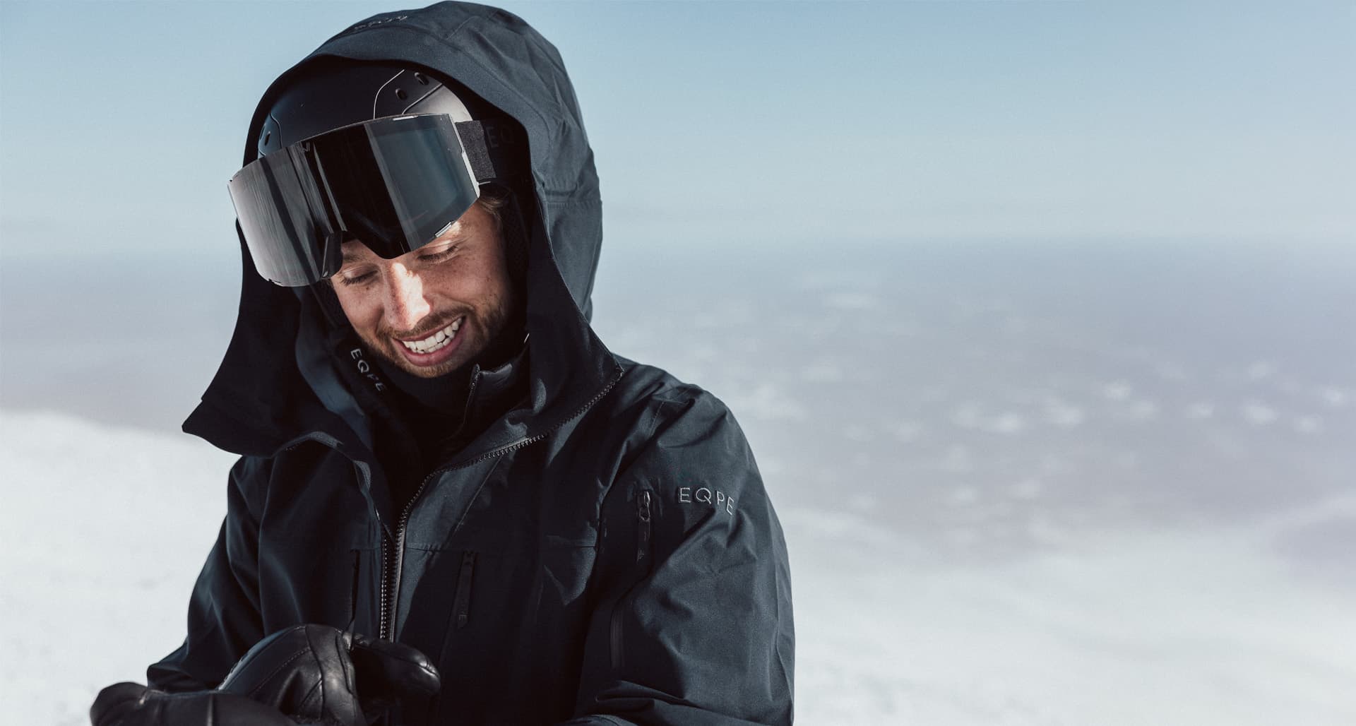 A person in a black winter jacket and goggles smiles while adjusting their gloves in a snowy landscape.