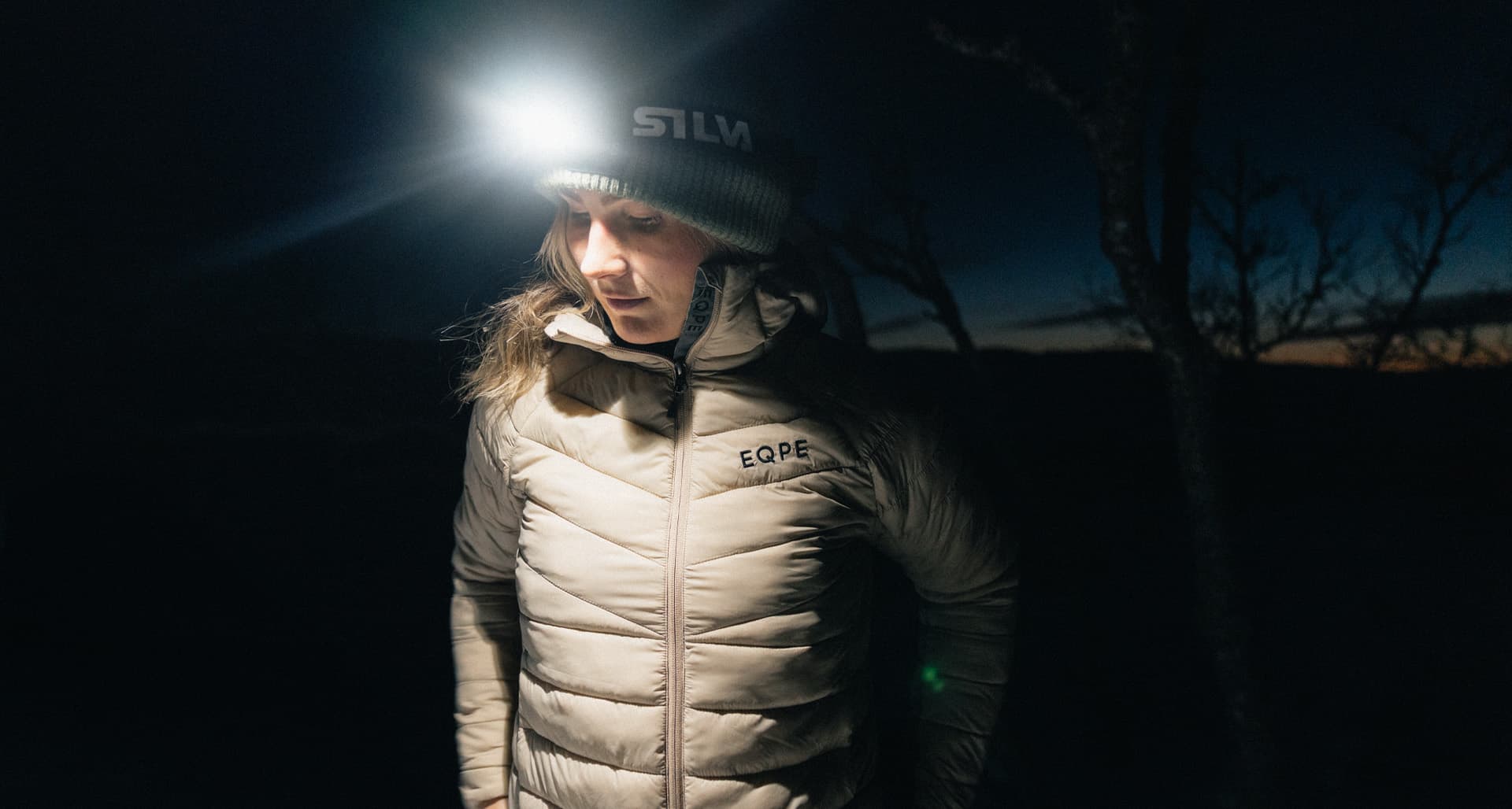 A person wearing a headlamp and a warm jacket stands outdoors in the dark, with a faint evening sky in the background.