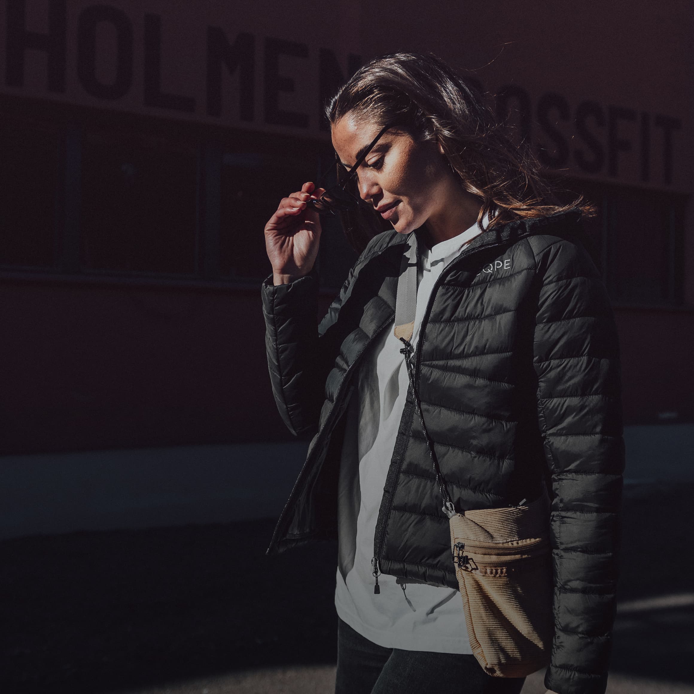 Woman in a black jacket and sunglasses outside, standing against a dark background with "HOLMEN CROSSFIT" partially visible.