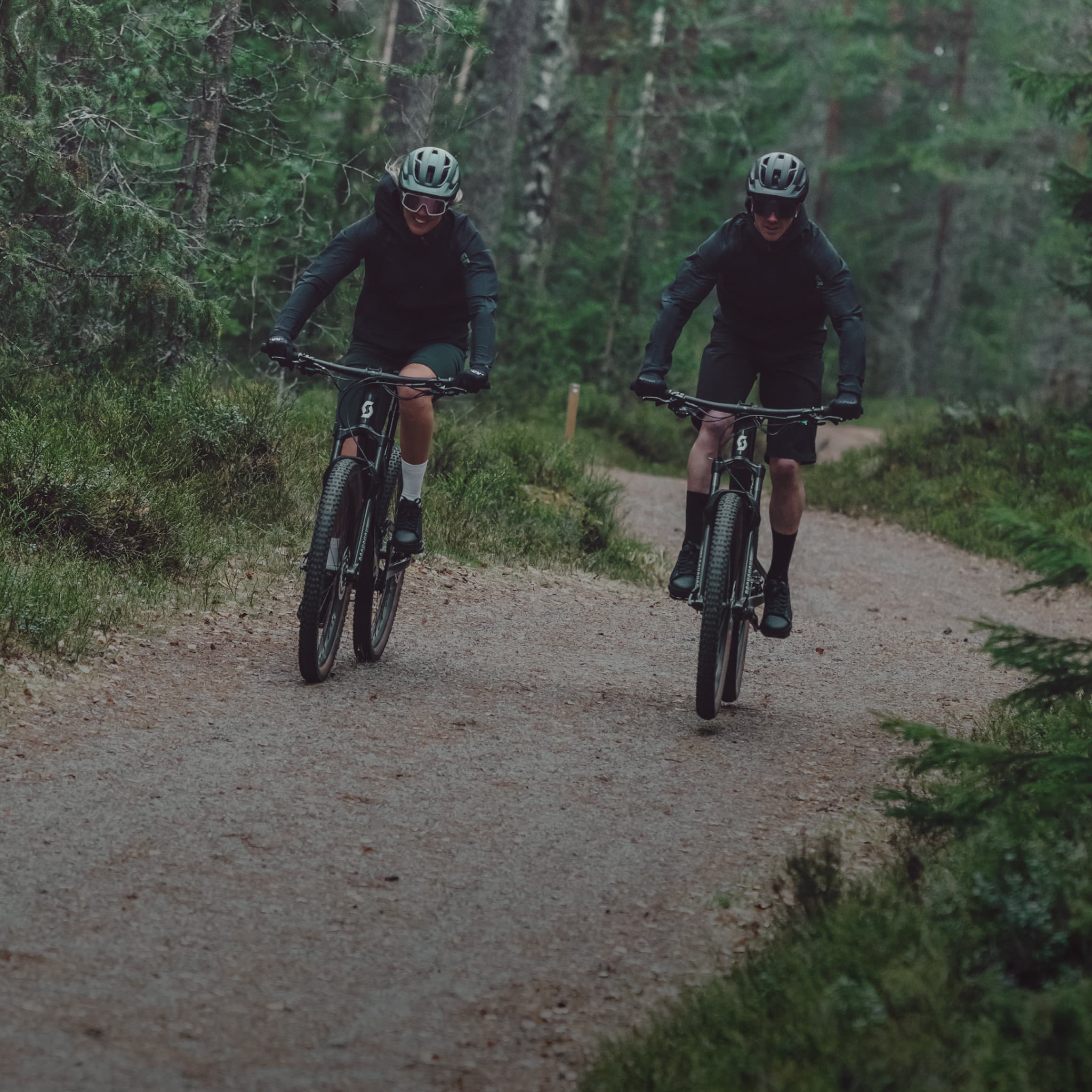 Two cyclists on mountain bikes ride along a dirt path in a lush forest, wearing dark clothing and helmets.
