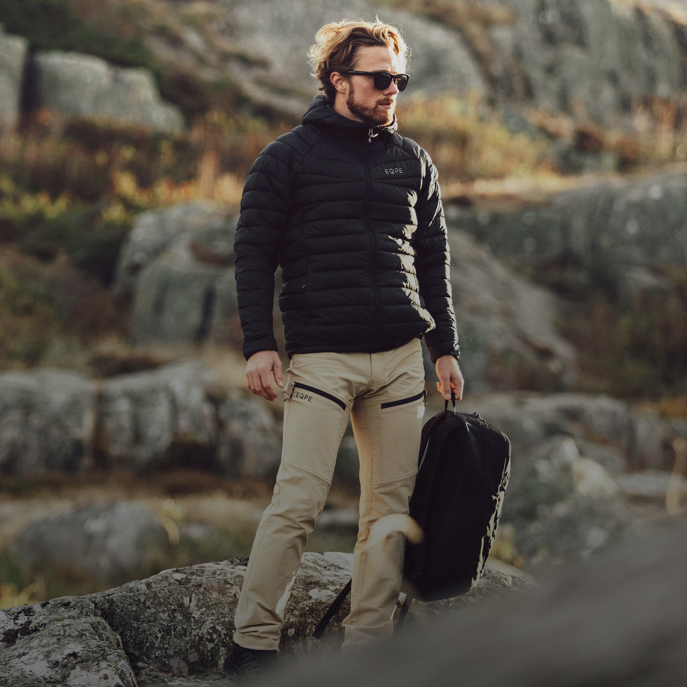 Man in a black jacket and beige pants stands on rocky terrain, holding a black backpack, with sunlight illuminating the background.