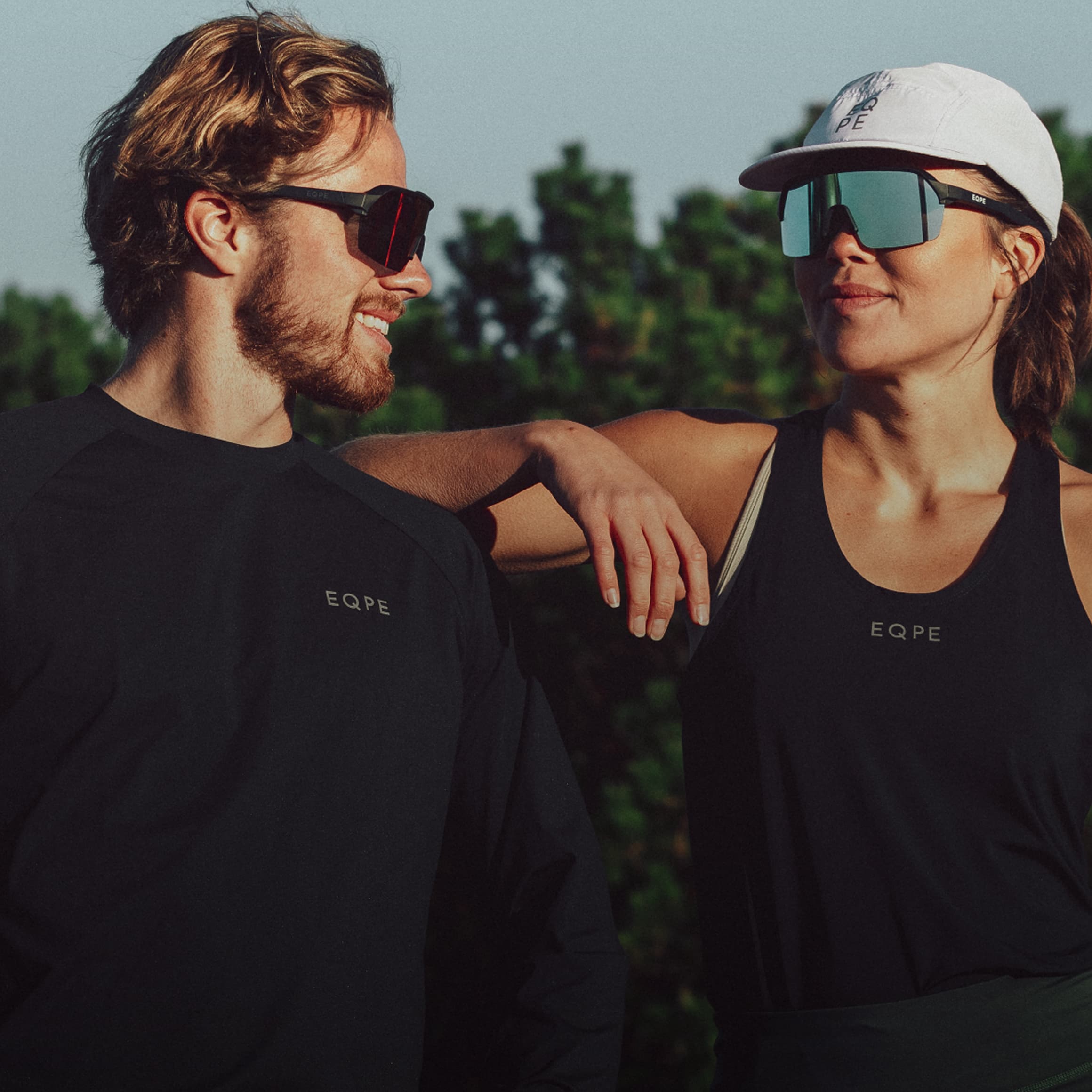 Two people wearing sunglasses and EQPE activewear smile at each other outdoors, with greenery in the background.