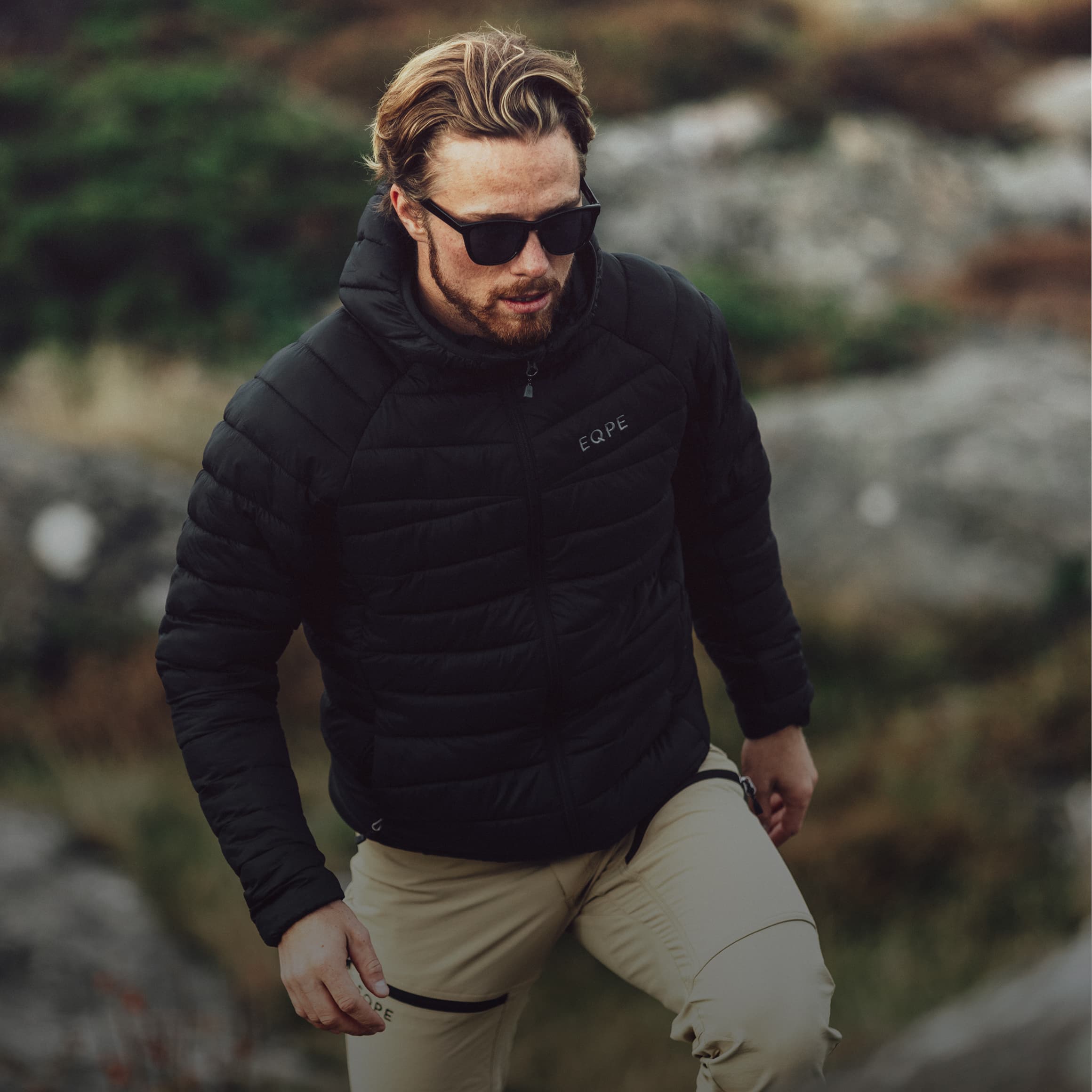 Man in sunglasses and a black puffer jacket walking outdoors on a rocky terrain with blurred greenery in the background.