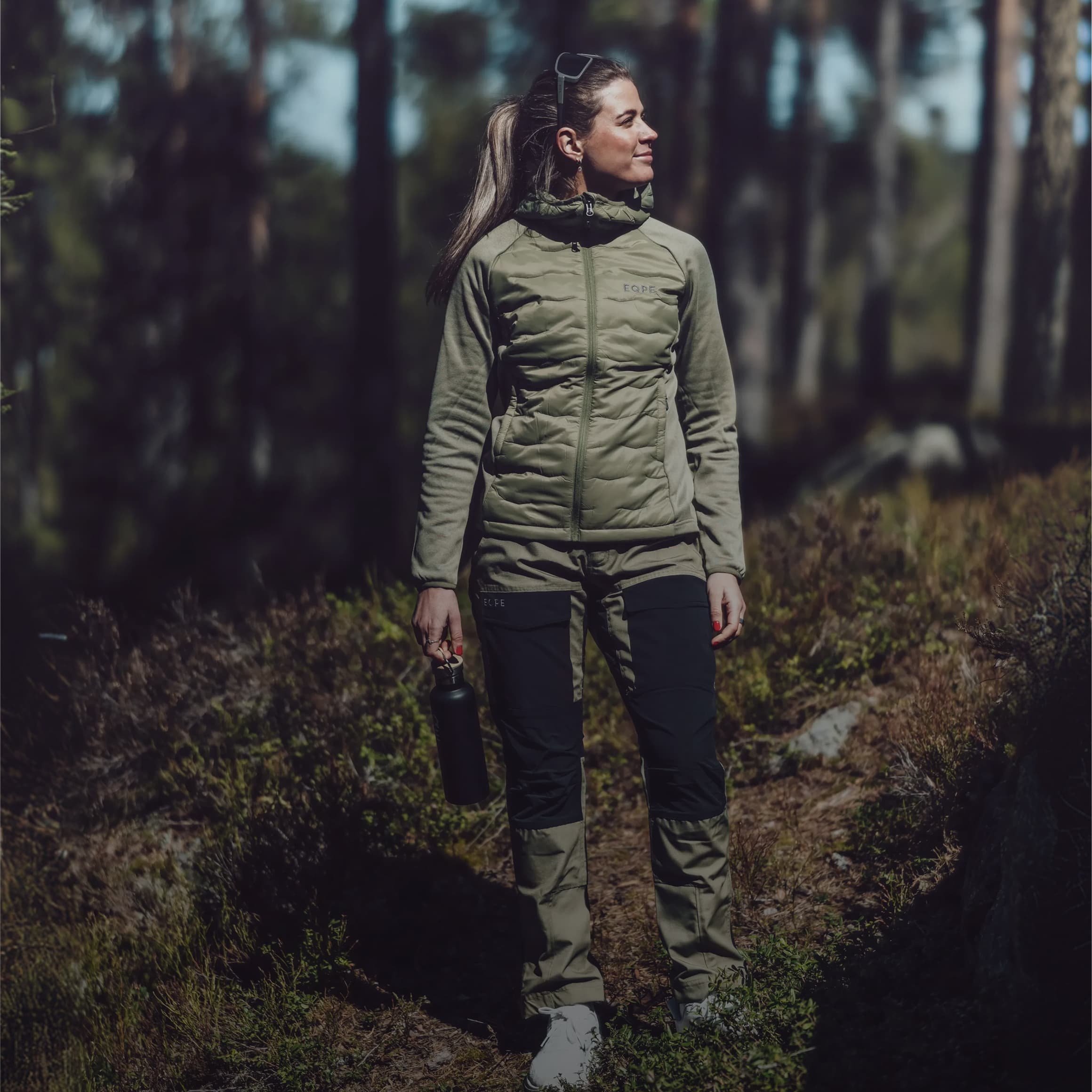 A woman in outdoor gear stands in a sunlit forest holding a bottle, looking to the side.