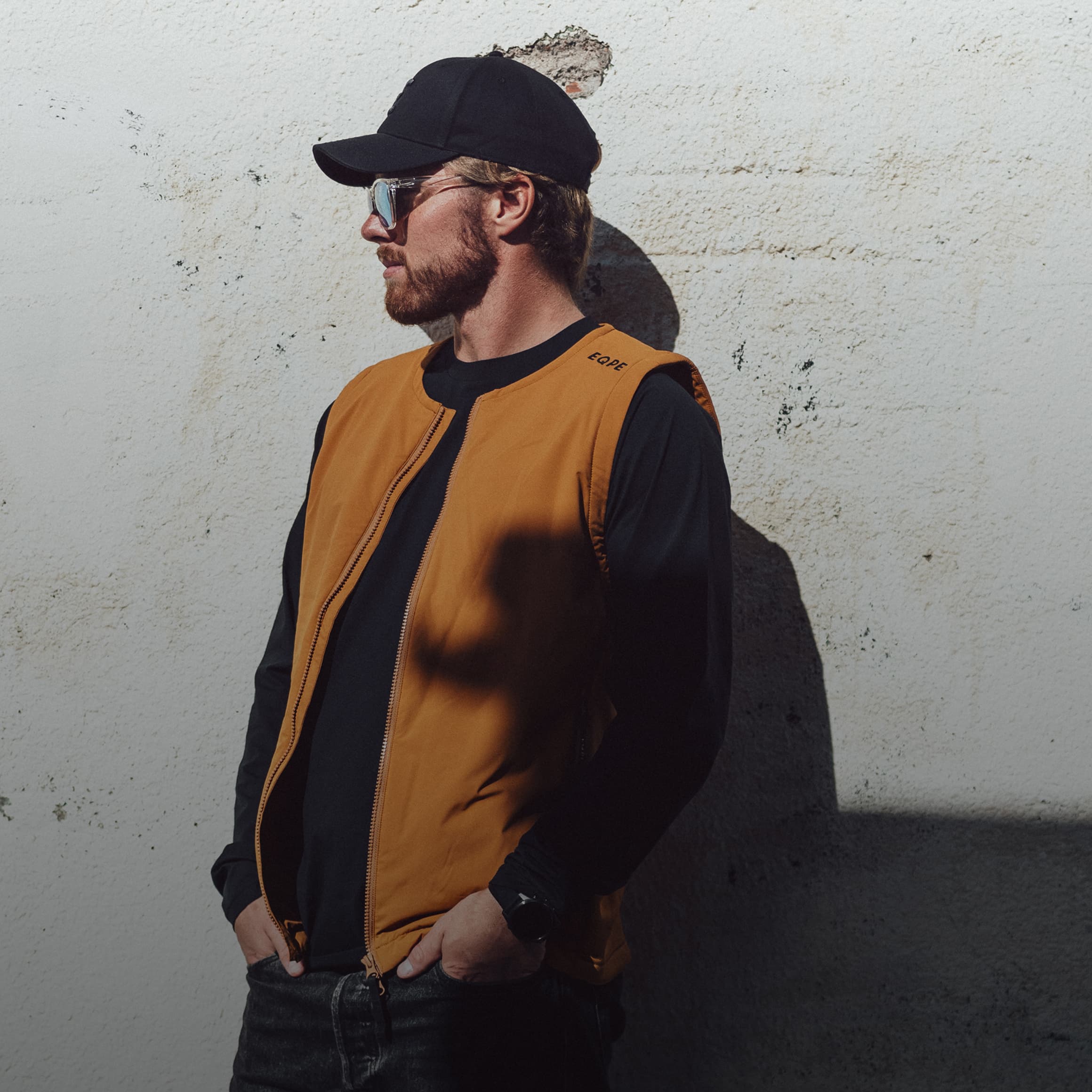 Man in a black cap and sunglasses wearing an orange vest, standing against a textured white wall in sunlight.