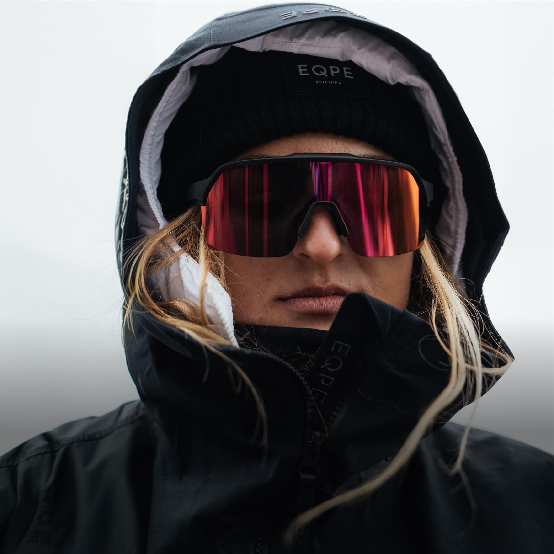 Person in a black hooded jacket and beanie, wearing reflective red ski goggles, with long hair, against a neutral background.
