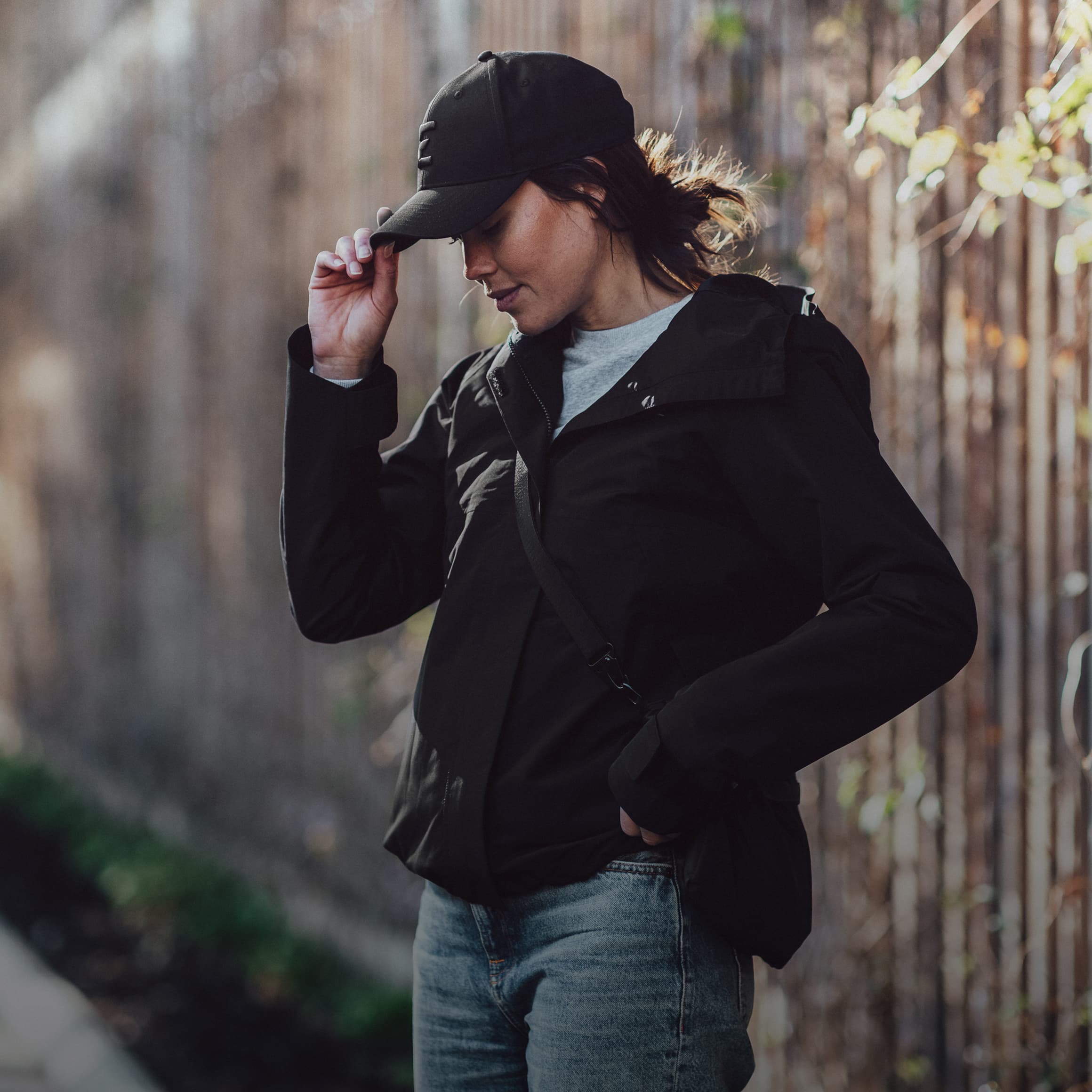 Person in a black jacket and cap walking outdoors, adjusting cap brim, with a softly blurred background of a wooden fence and foliage.