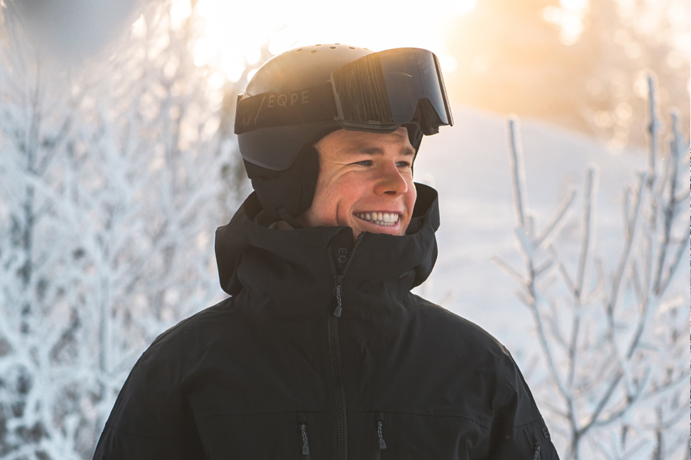 A person in a black ski jacket and helmet smiles while standing in a snowy landscape.