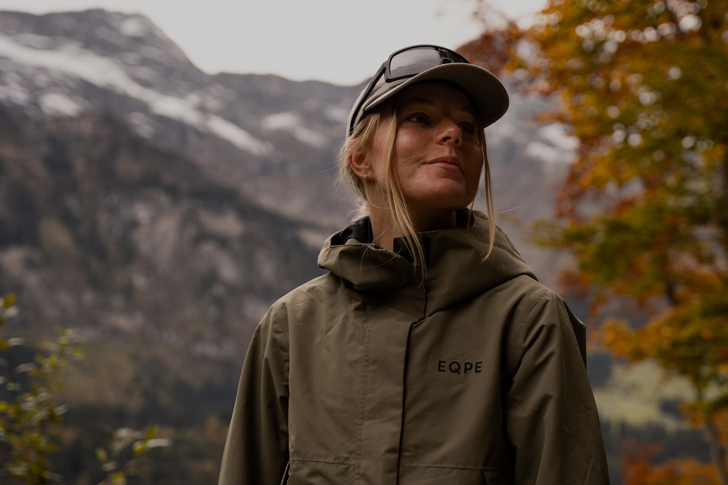 Woman in a green jacket and cap stands outdoors, with autumn trees and snow-capped mountains in the background.
