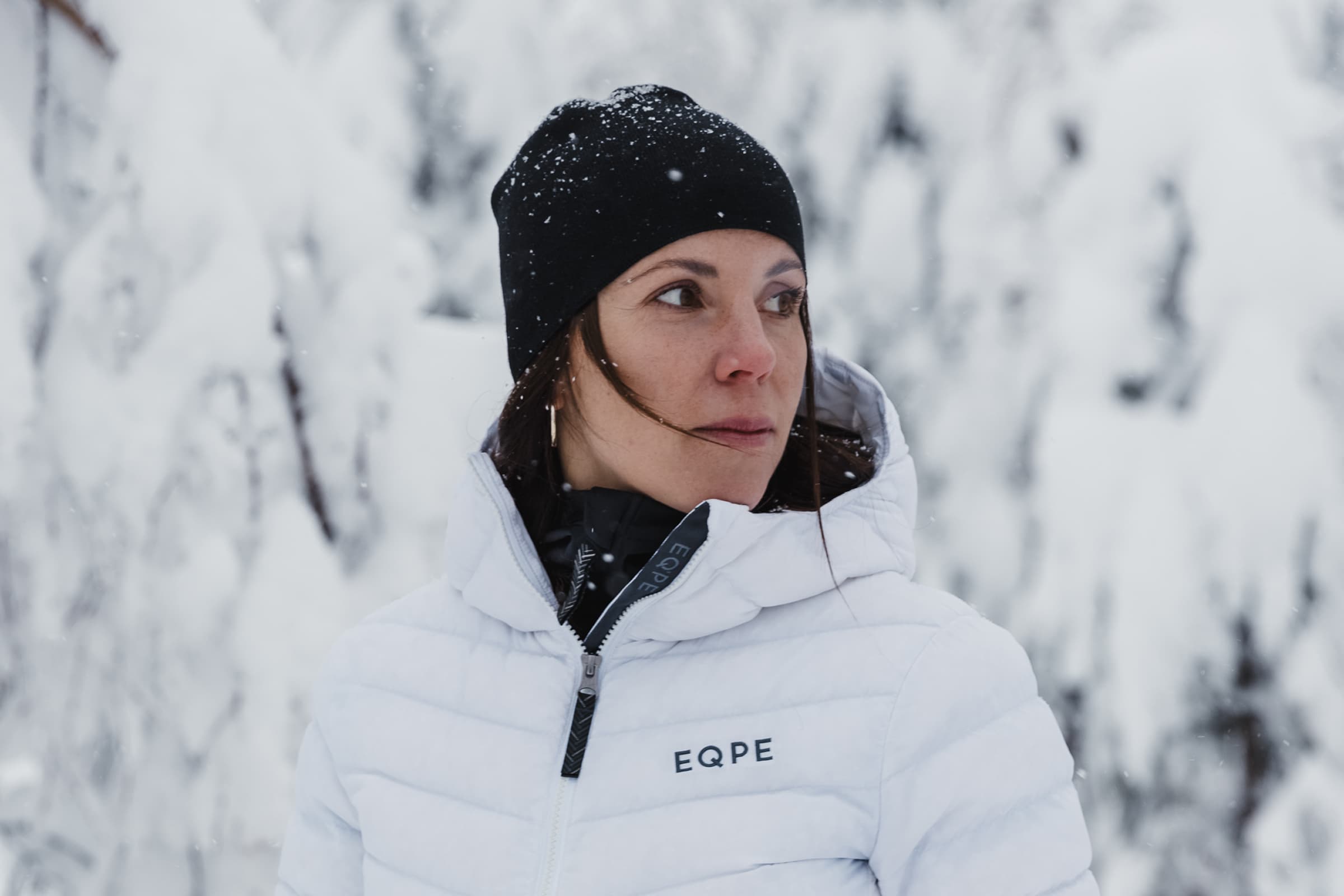 Woman in a white winter jacket and black beanie standing in a snowy forest.
