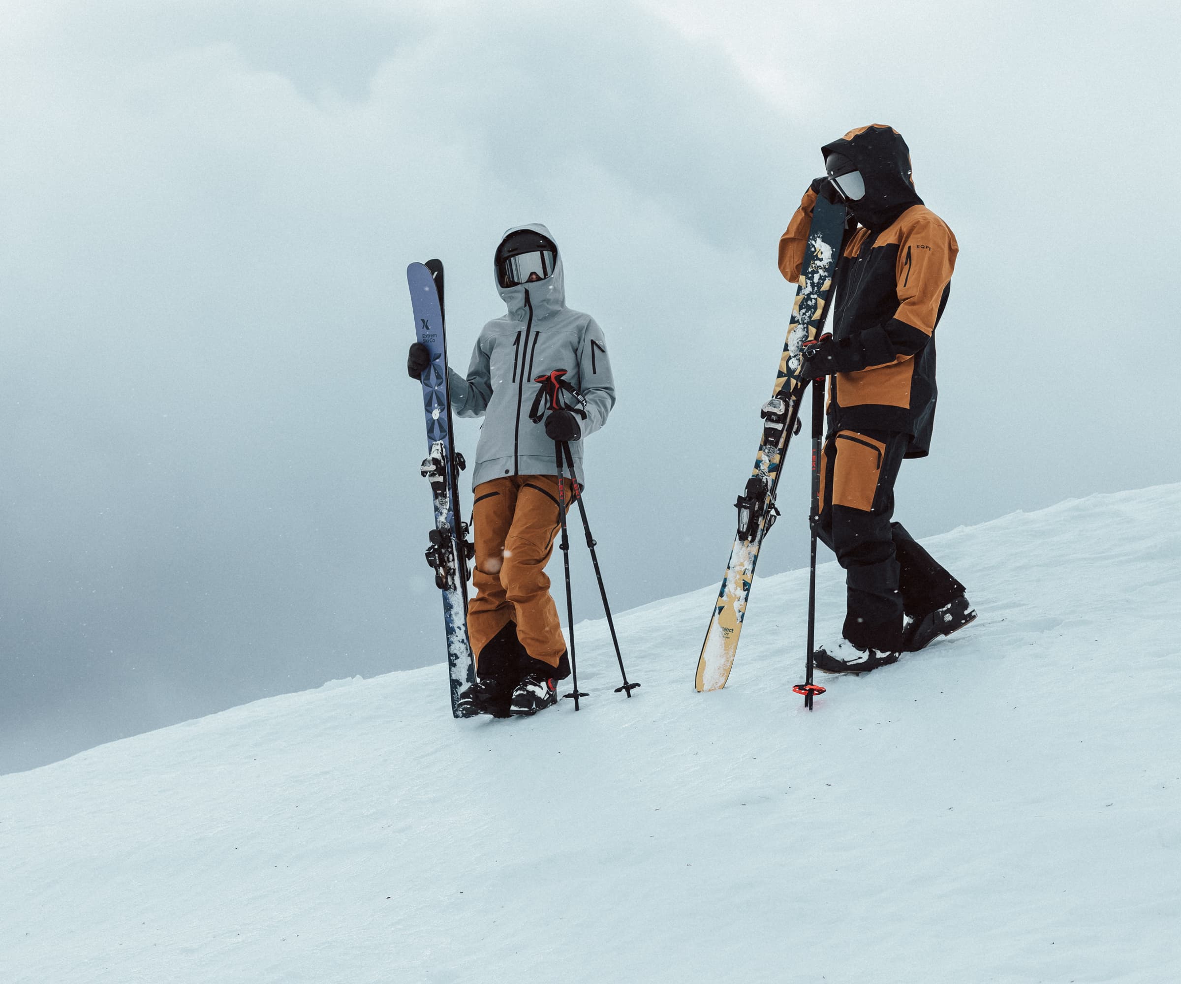 Two skiers in winter gear stand on a snowy slope, holding skis, with a cloudy sky in the background.