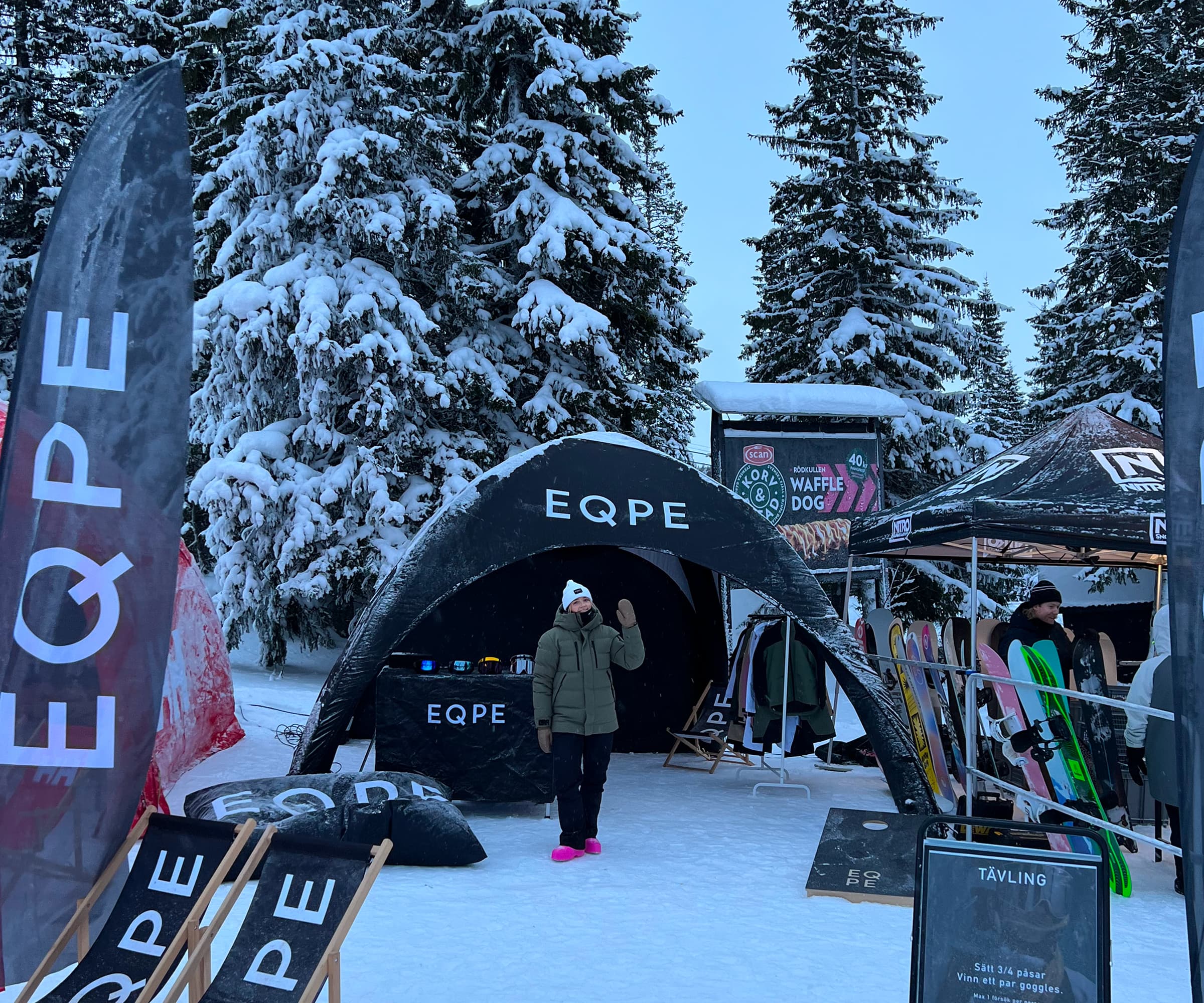Snowy outdoor event with a person in a winter jacket and hat standing near a black EQPE tent, surrounded by trees and ski equipment.