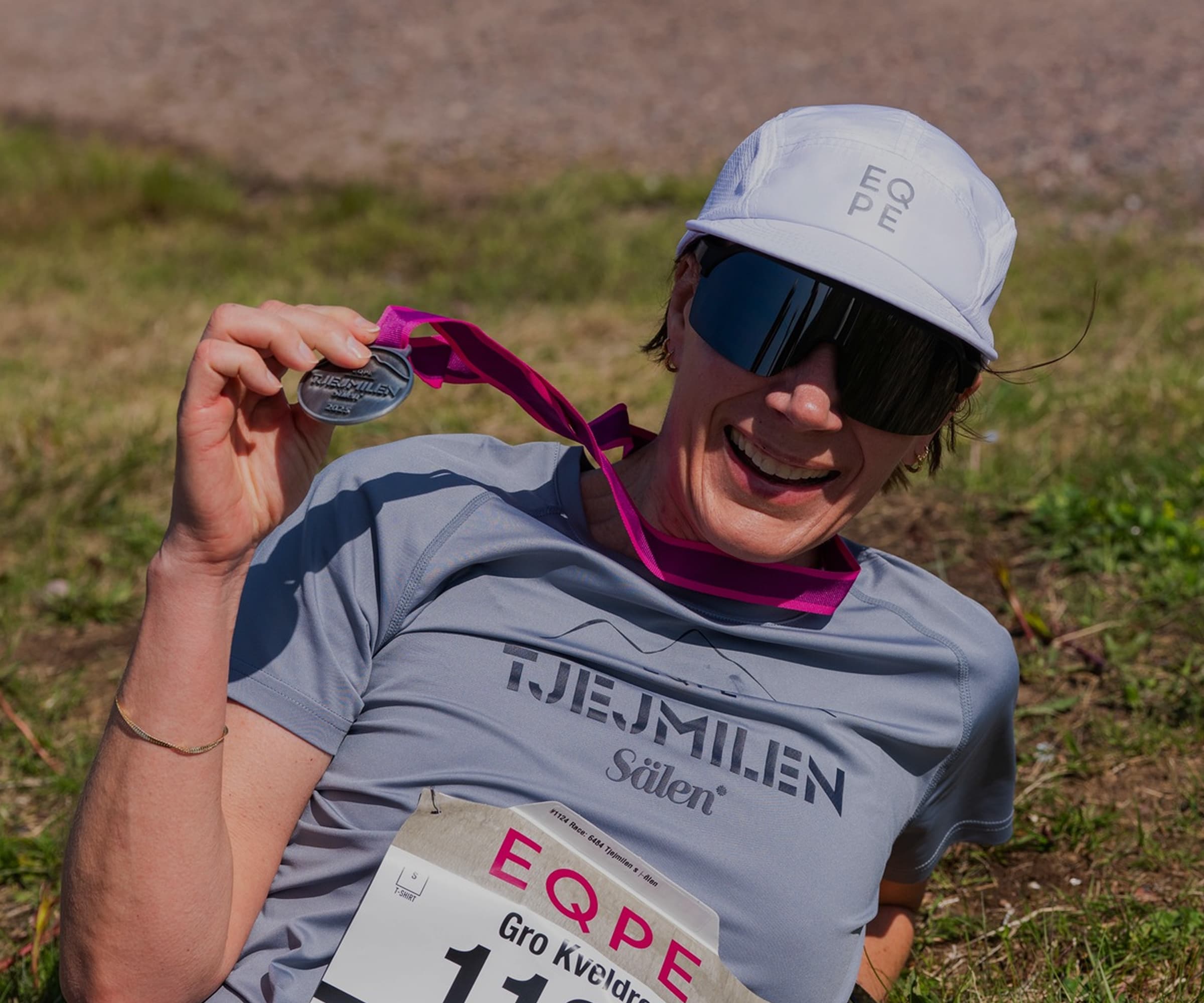 Smiling person in sunglasses and cap holds a medal.