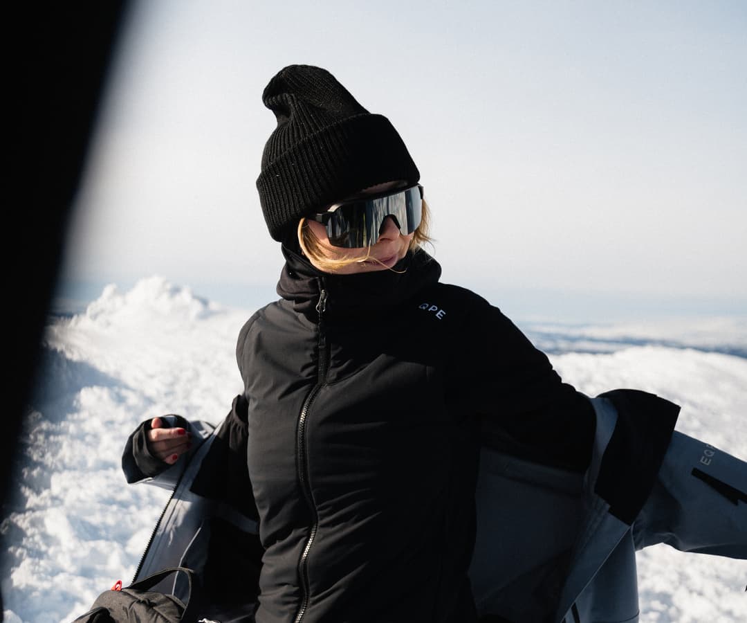 Person in black winter gear standing on a snowy landscape, adjusting their jacket under a clear sky.