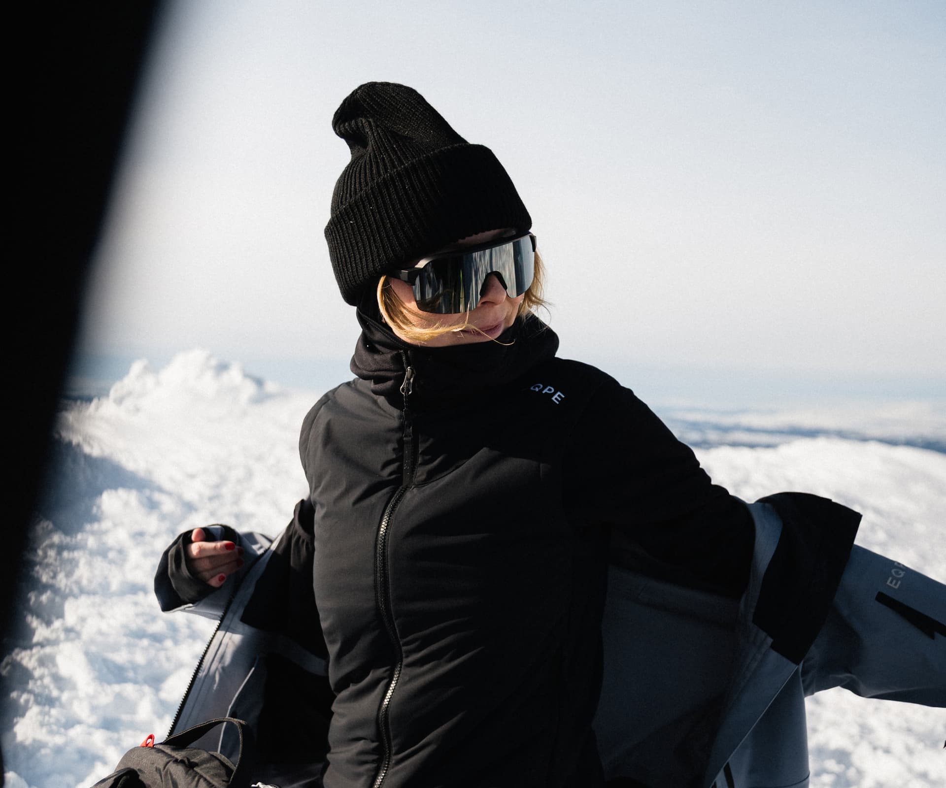 Person in black winter gear standing on a snowy landscape, adjusting their jacket under a clear sky.