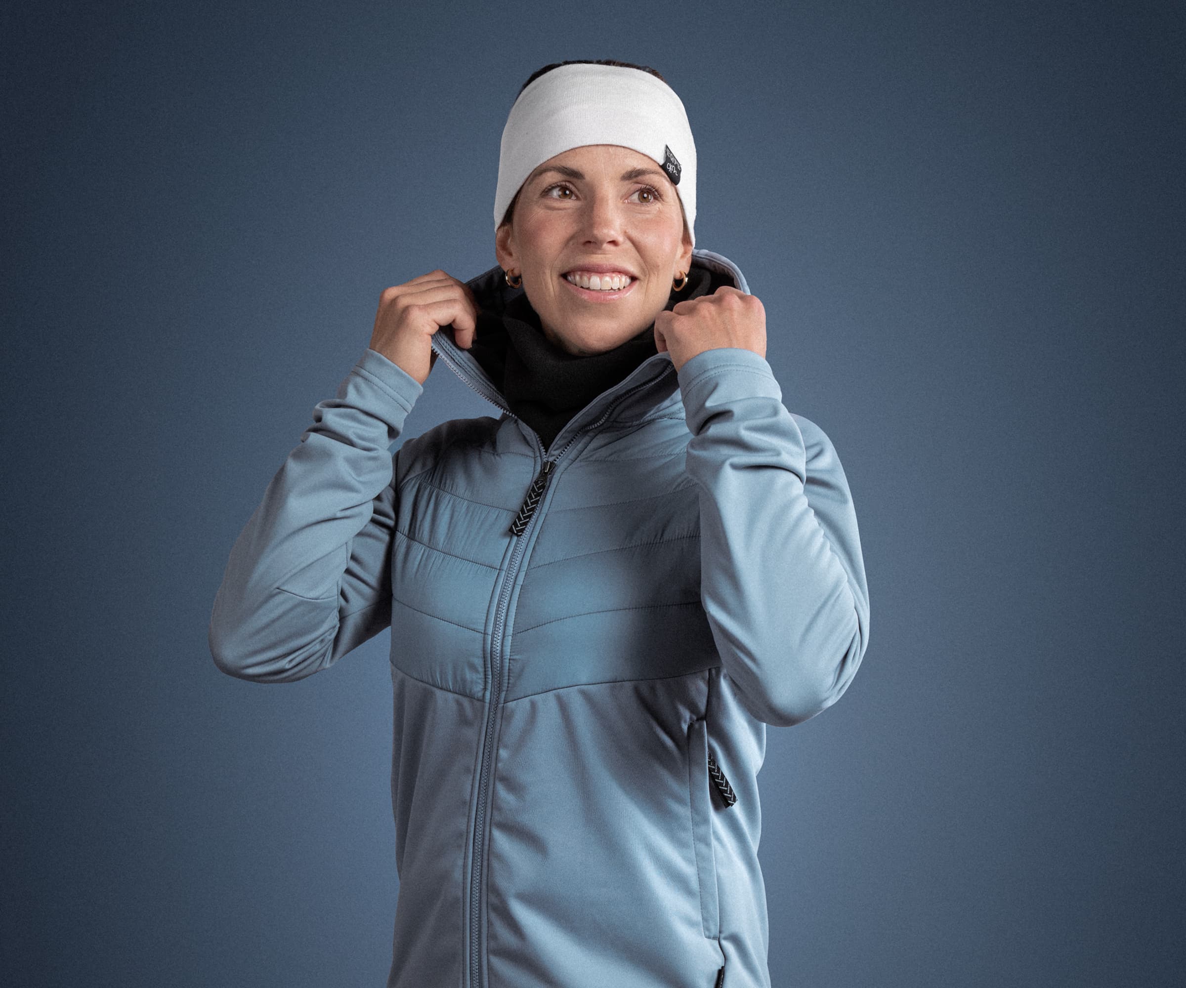 Person in a blue jacket and white headband smiling, adjusting their collar against a plain blue background.
