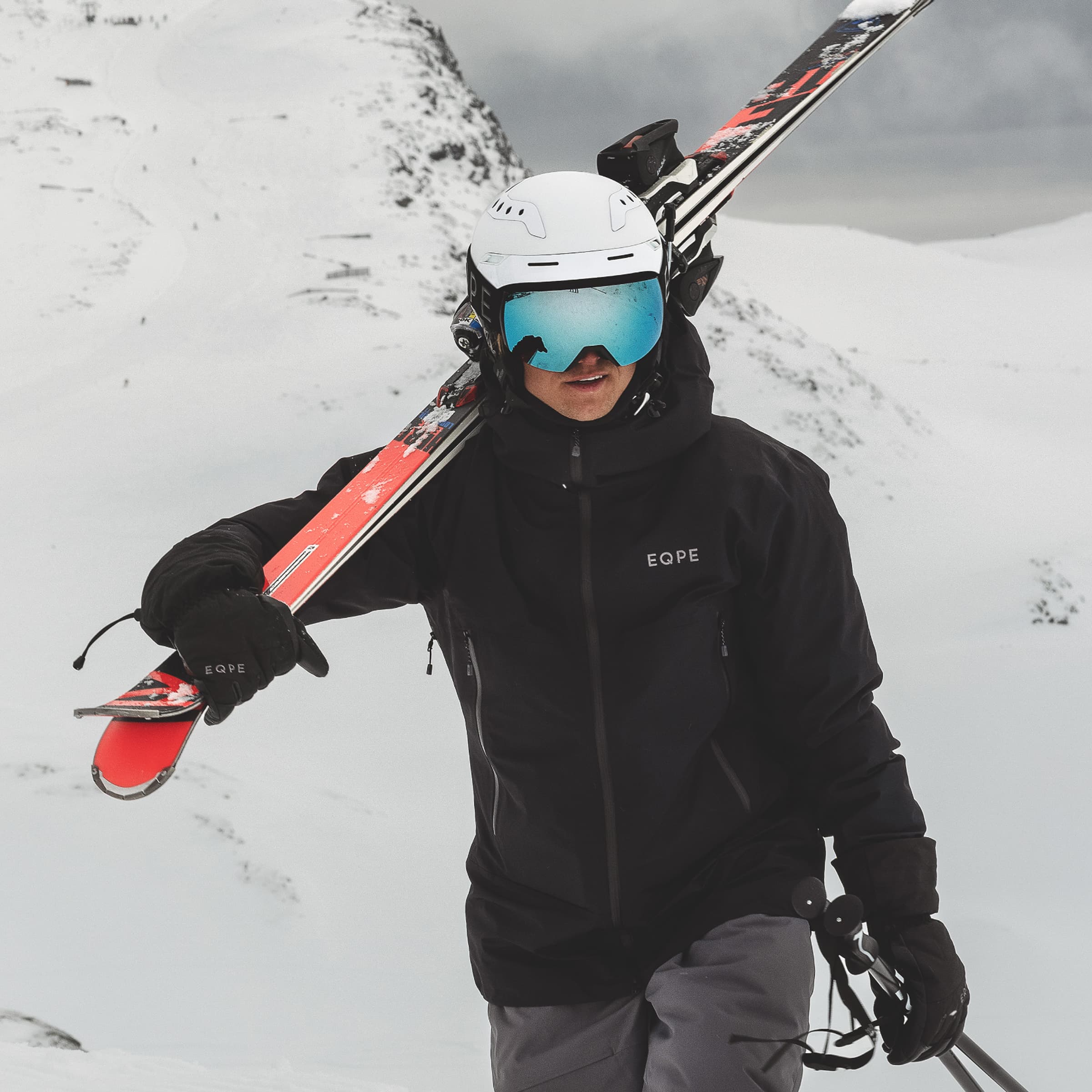 A skier in black ski gear carries skis on their shoulder, walking through a snowy mountain landscape.