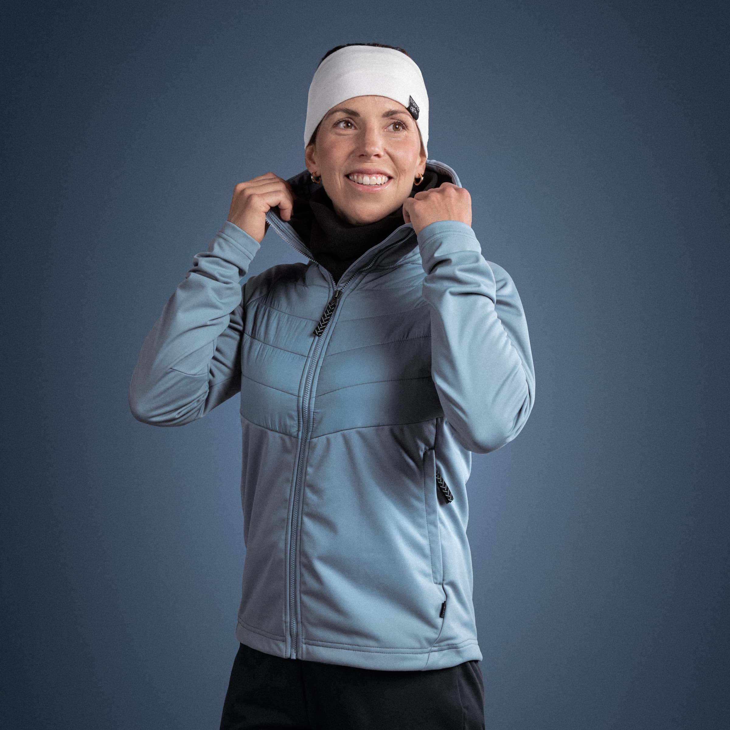 Person in a blue jacket and white headband smiling, adjusting their collar against a plain blue background.