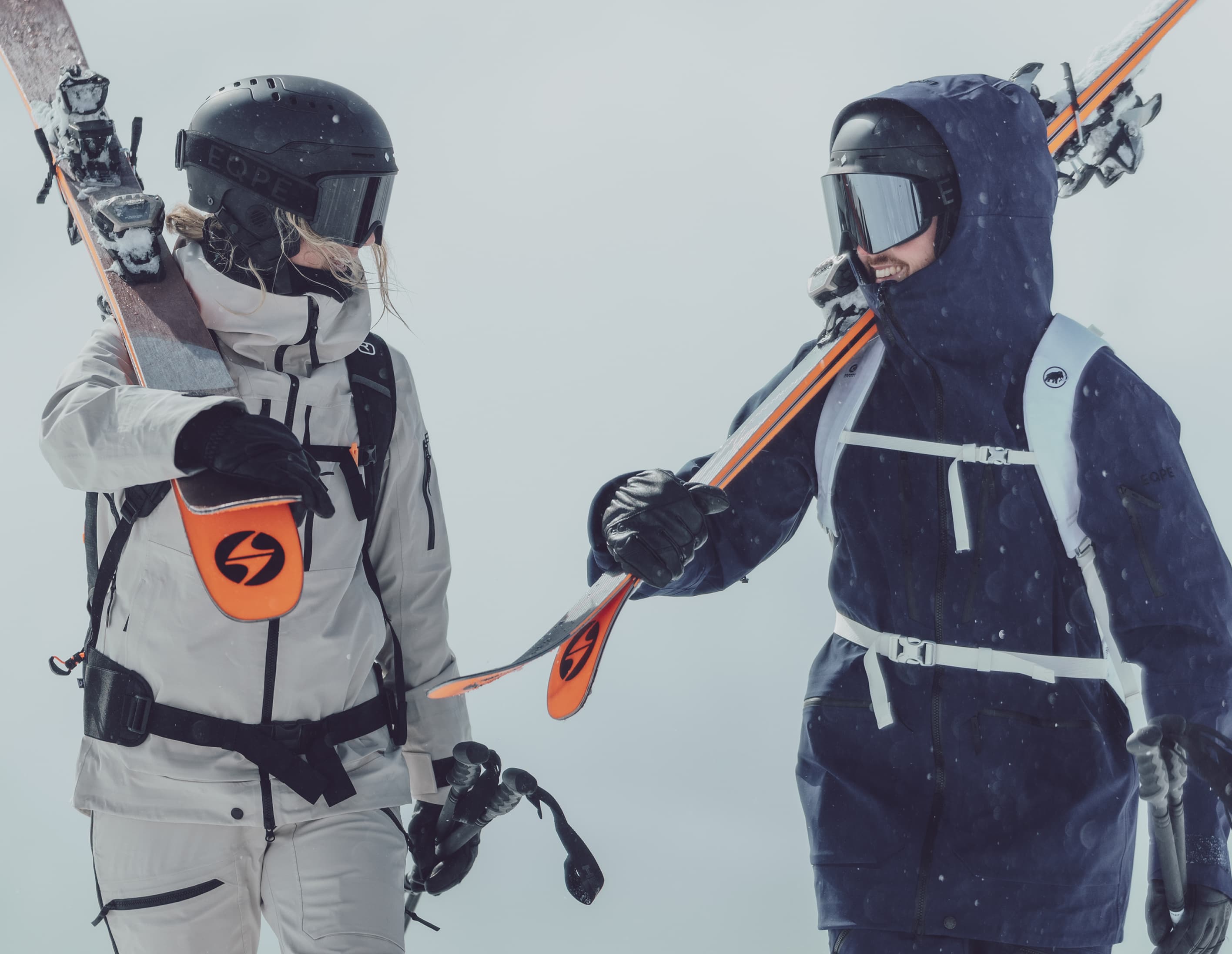 Two skiers in winter gear, holding skis over their shoulders, stand in snowy conditions, wearing helmets and goggles.