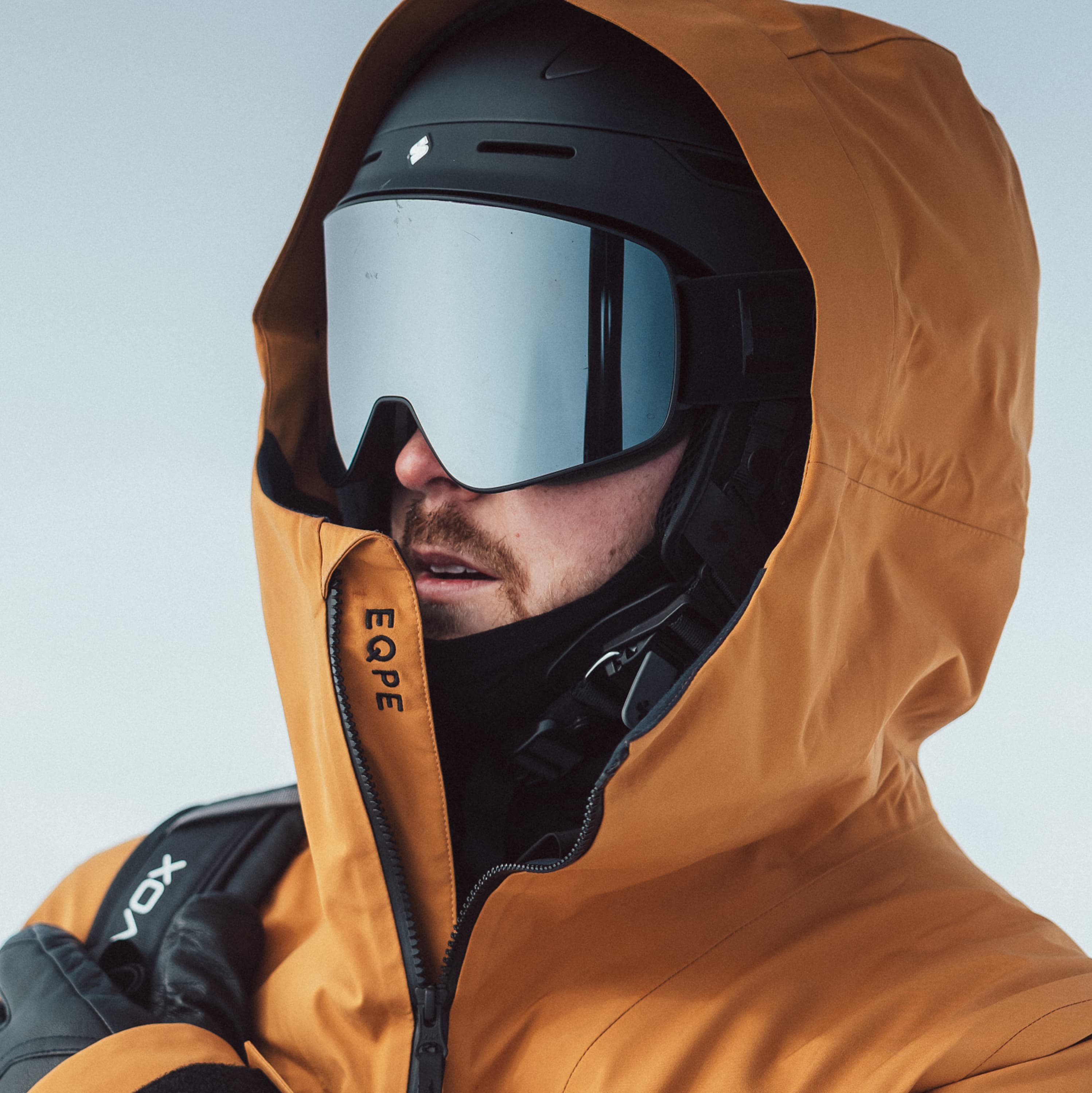Person in orange winter jacket and helmet, wearing reflective ski goggles, against a snowy backdrop.
