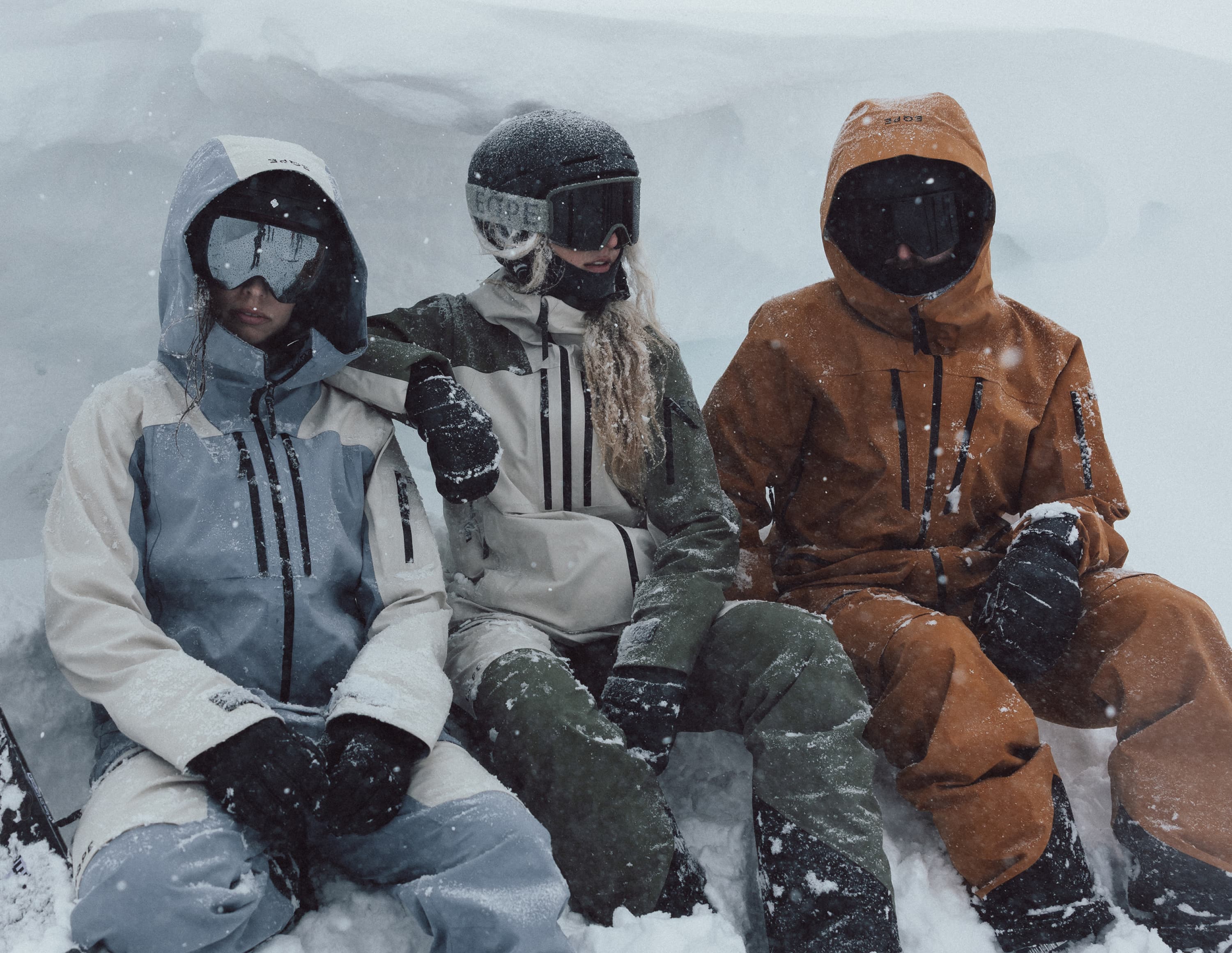 Three people in ski gear sit in a snowy landscape, surrounded by falling snow.