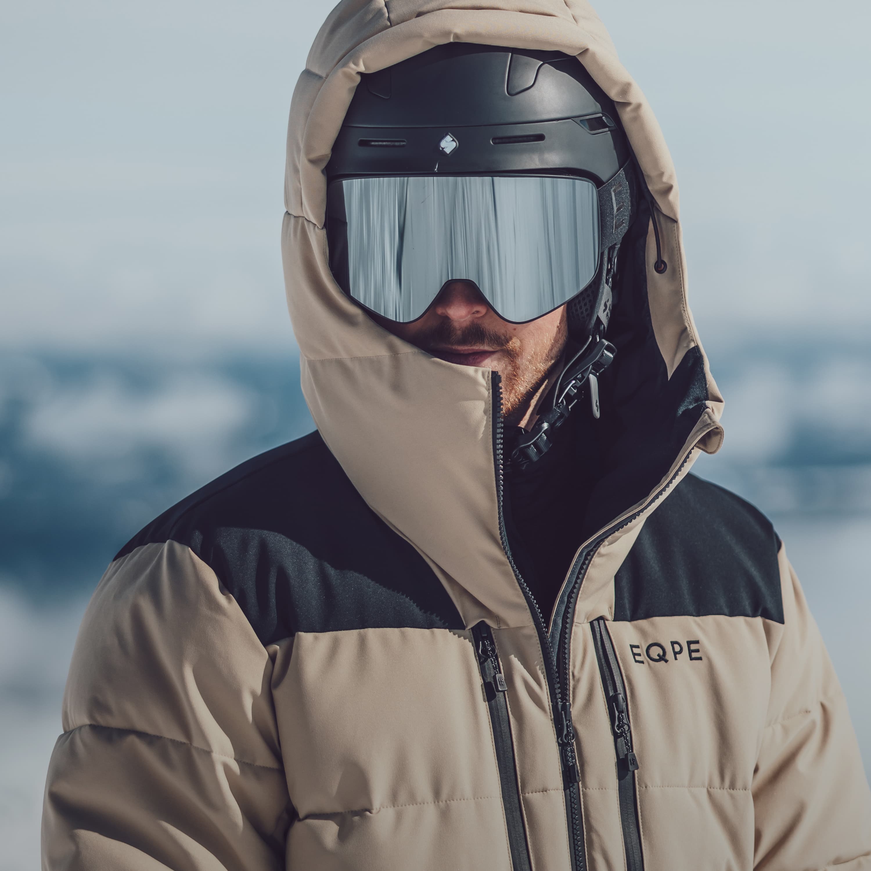 Person in a beige and black ski jacket and helmet with reflective goggles, standing outdoors in a snowy landscape.