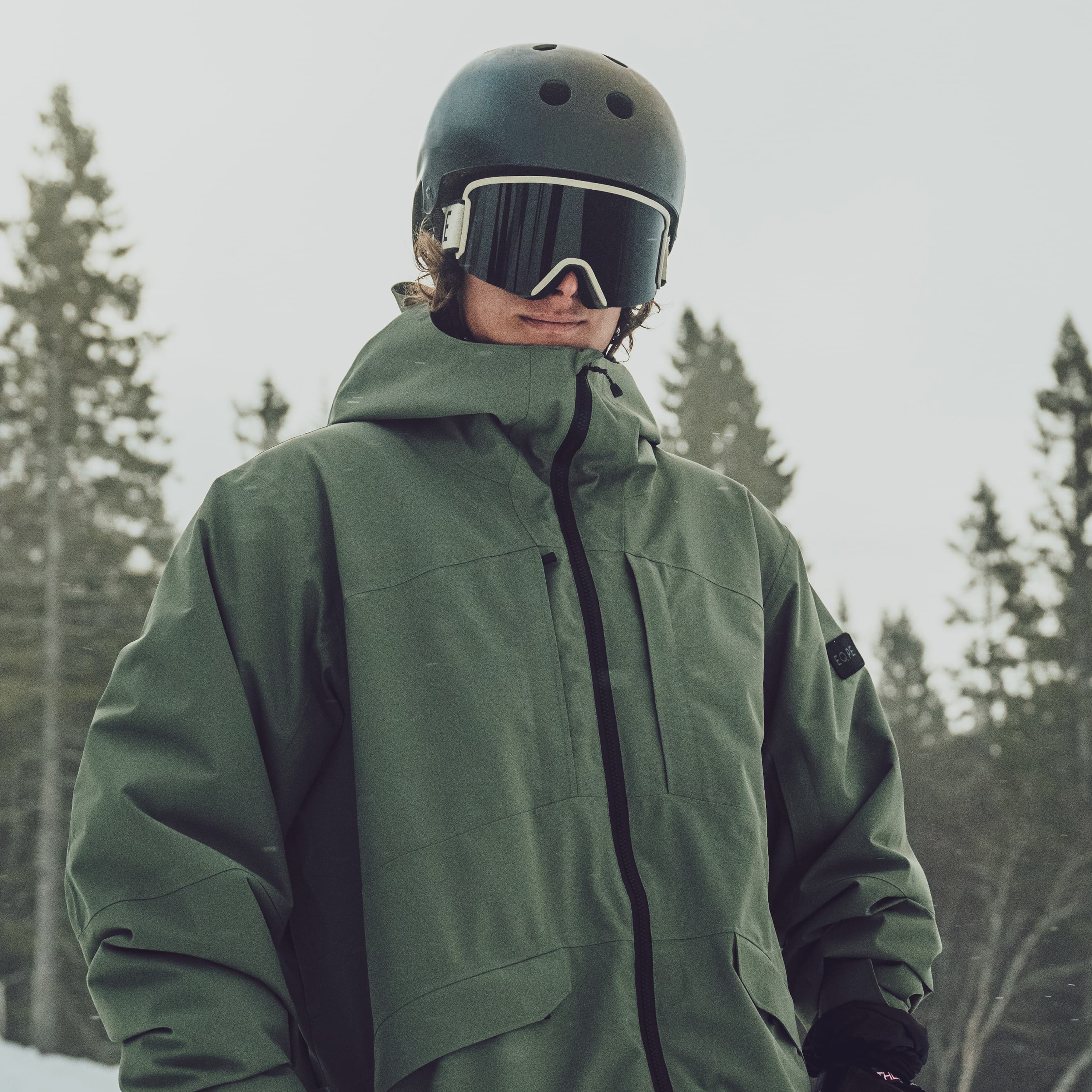 Person in a green ski jacket, standing outdoors with snow-covered trees in the background.