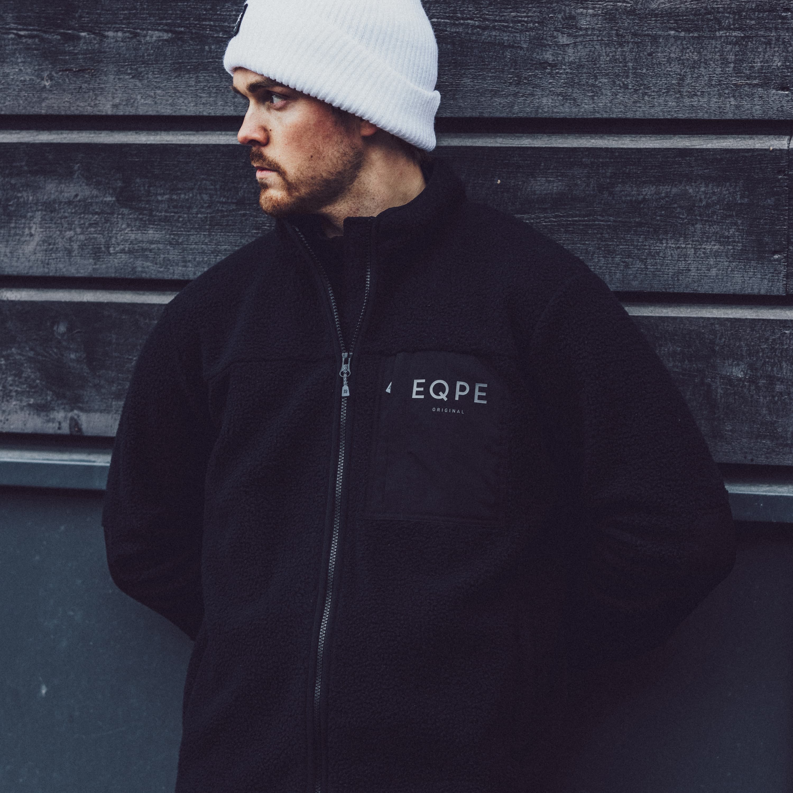 Man in a black jacket and white beanie stands against a dark wooden wall, looking to the side.