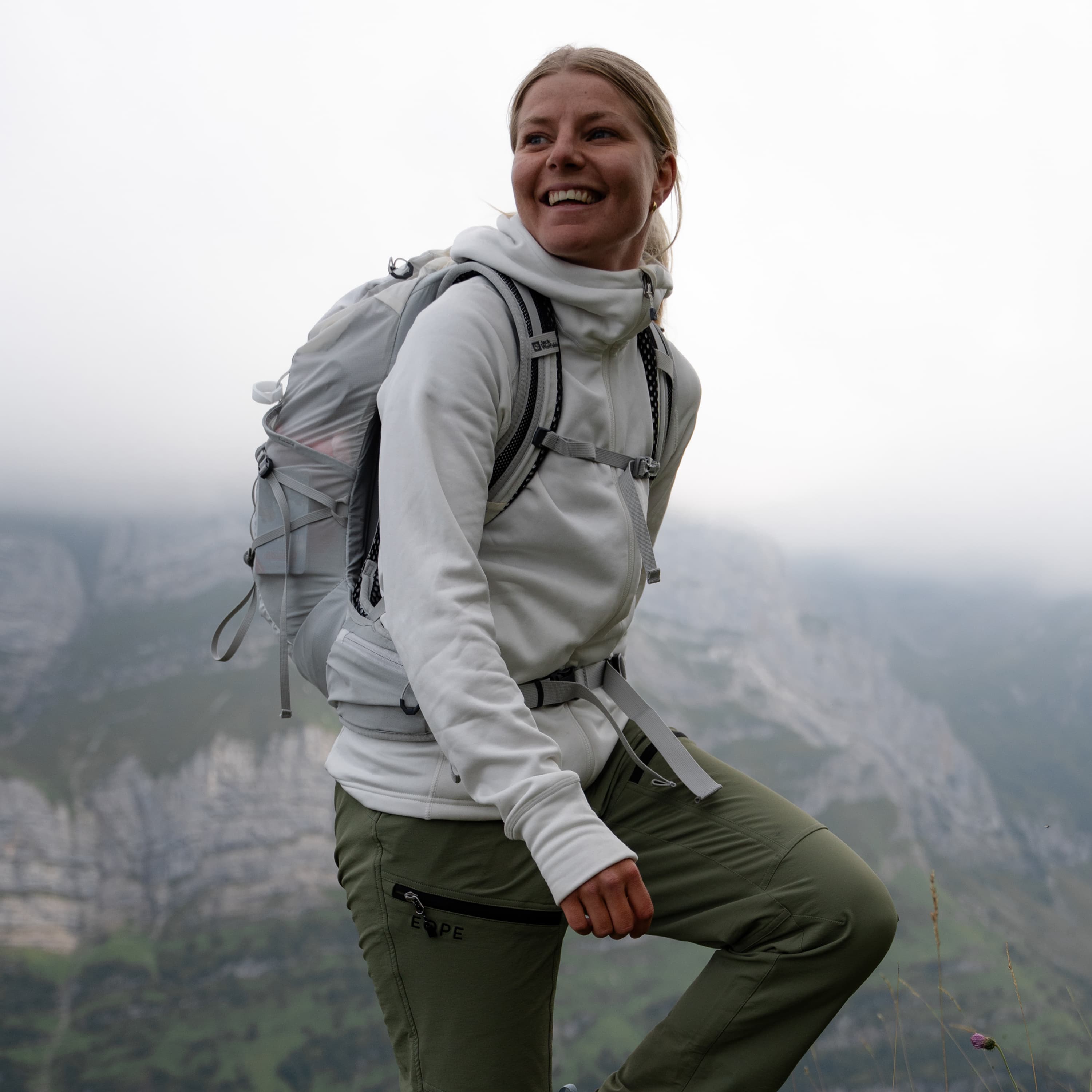 Smiling woman in a white hoodie and green pants hiking.