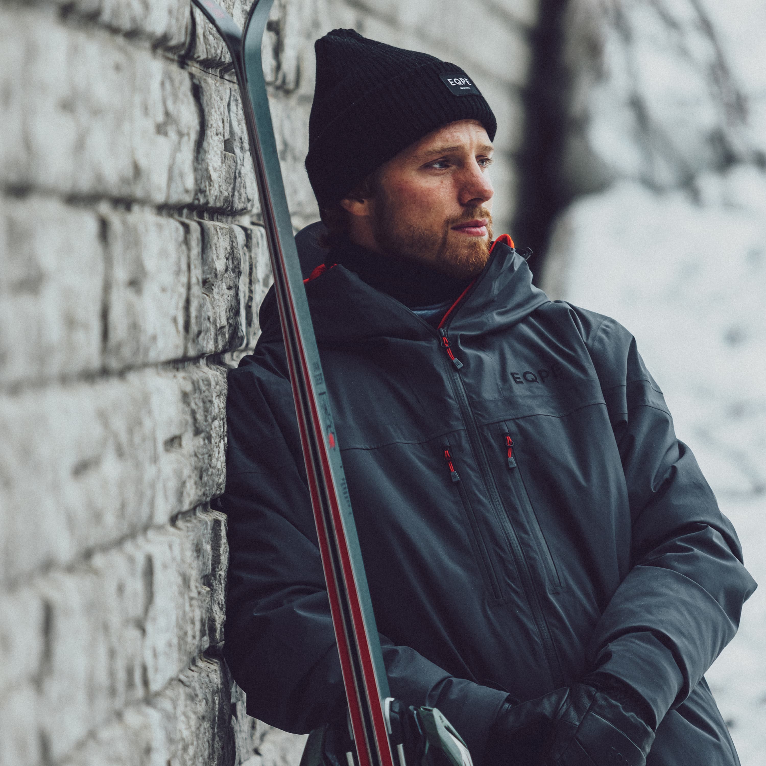Man in winter attire leaning against a stone wall, holding skis. Snowy background, wearing a beanie and gloves, looking into the distance.