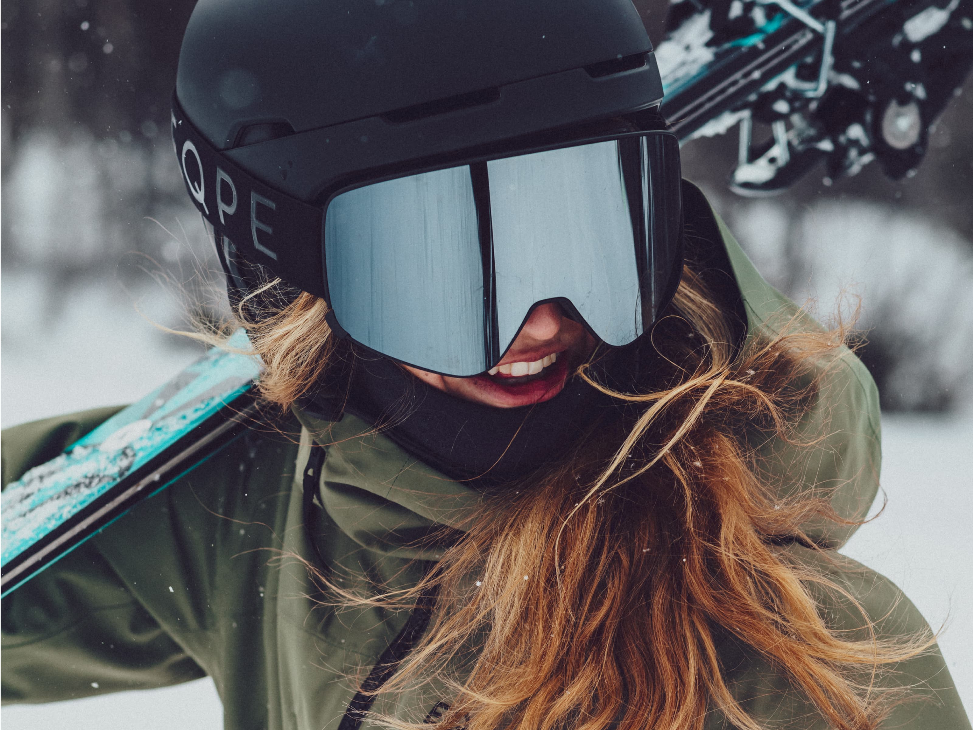Person in a green jacket and helmet smiling, carrying skis on their shoulder in a snowy setting.