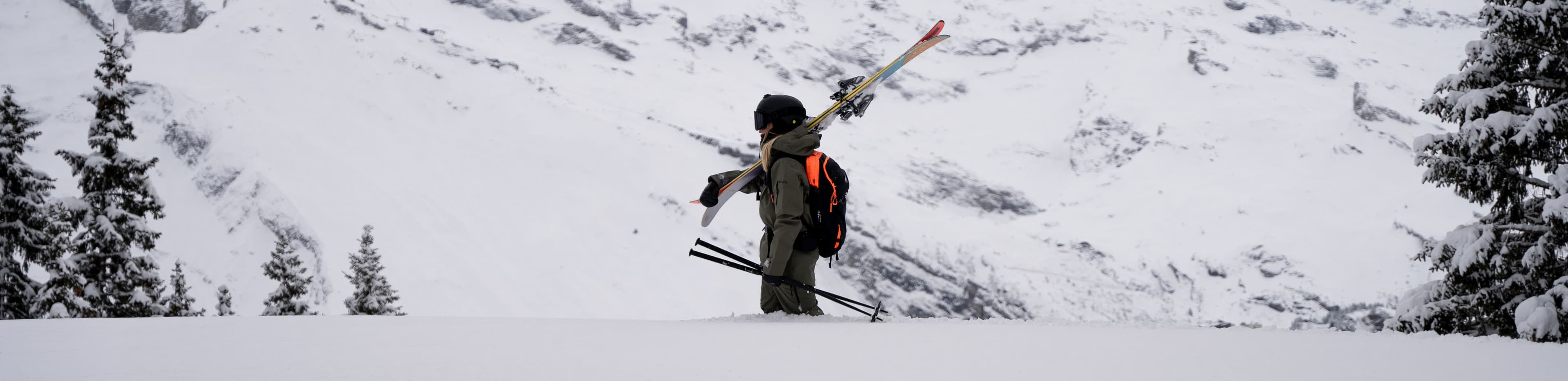 Skier in snowy landscape, carrying skis and poles on their shoulder, surrounded by snow-covered trees and mountains.